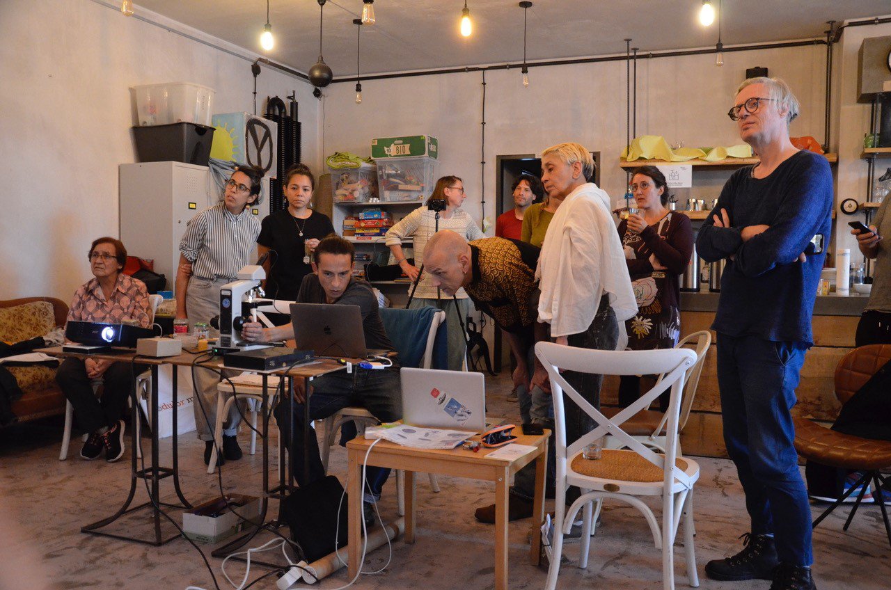 Das Foto zeigt einen Workshop-Raum. In der Mitte kümmert sich eine Person sitzend um den Beamer. Drumherum stehen 10 weitere Personen, die fast alle Richtung Leinwand gucken. Sie haben zum Teil Getränke in der Hand.