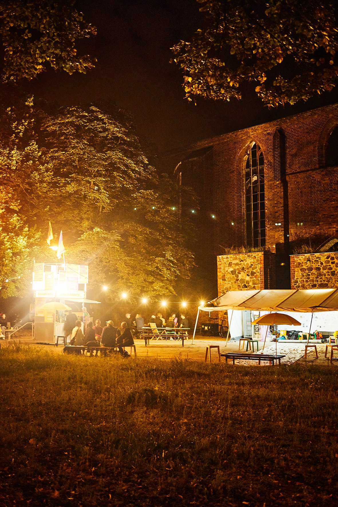 Es ist ein großer Platz vor einer Kirche bei nacht zu sehen. Links ist das Kioskgerüst. hell erleuchtet von Girlanden und Scheinwerfern. Leute sitzen auf Bierbänken um den Kiosk. Rechts ist ein Zelt aufgebaut, darunter ein Teppich und eine Sitzecke.