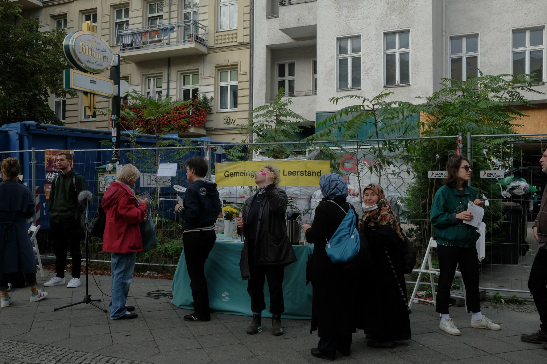 Es stehen 9 Personen zusammen, die sich zum Teil unterhalten. Am Bauzaun dahinter steht auf einem gelben Banner „Gemeingut statt Leerstand“. Hinterm Zaun liegt Baumaterial und ein großer blauer Baucontainer ist vor einem leergeräumten Haus aufgestellt.
