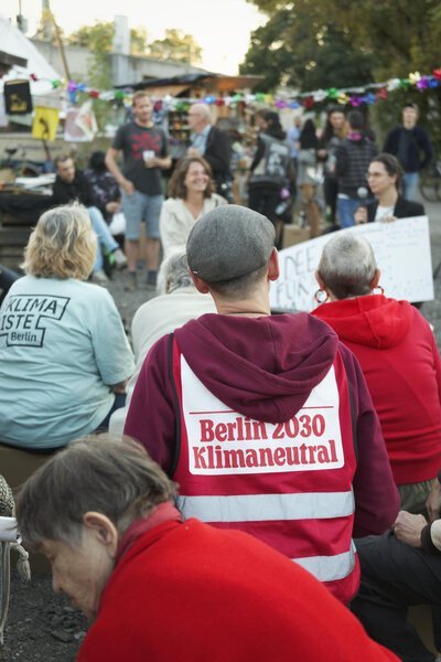 Menschen sitzen auf einem Platz, im Hintergrund ist eine Girlande gespannt. Im Vordergrund sitzt eine Person mit Hut, sie trägt eine Weste auf der steht: Berlin 2030 Klimaneutral