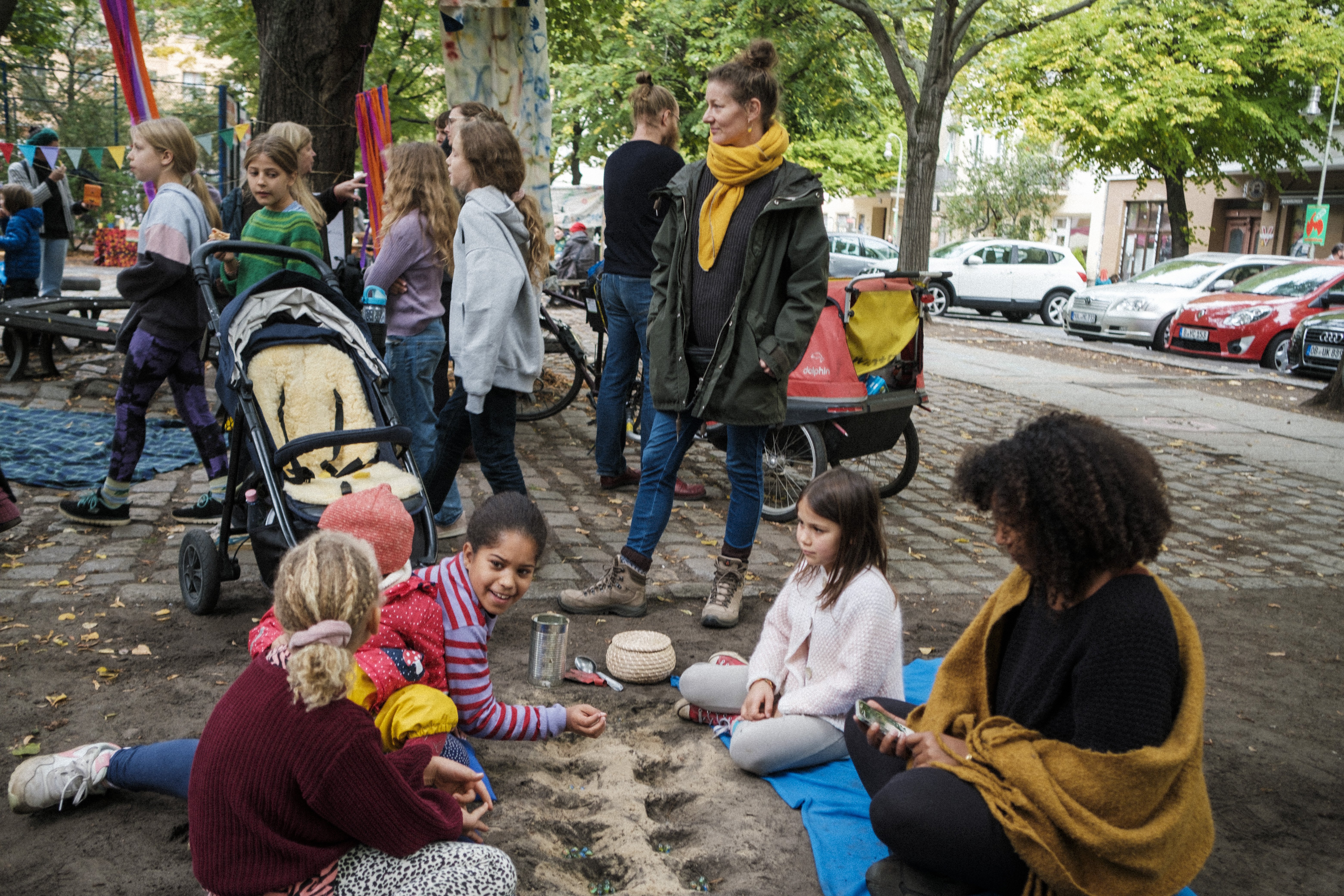 Im Vordergrund sitzen 4 Kinder mit einer erwachsenen Frau um ein in den Boden gegrabenes Spielfeld. Im Hintergrund läuft eine Gruppe Kinder vorbei und es stehen Erwachsenen mit Kinderwagen.