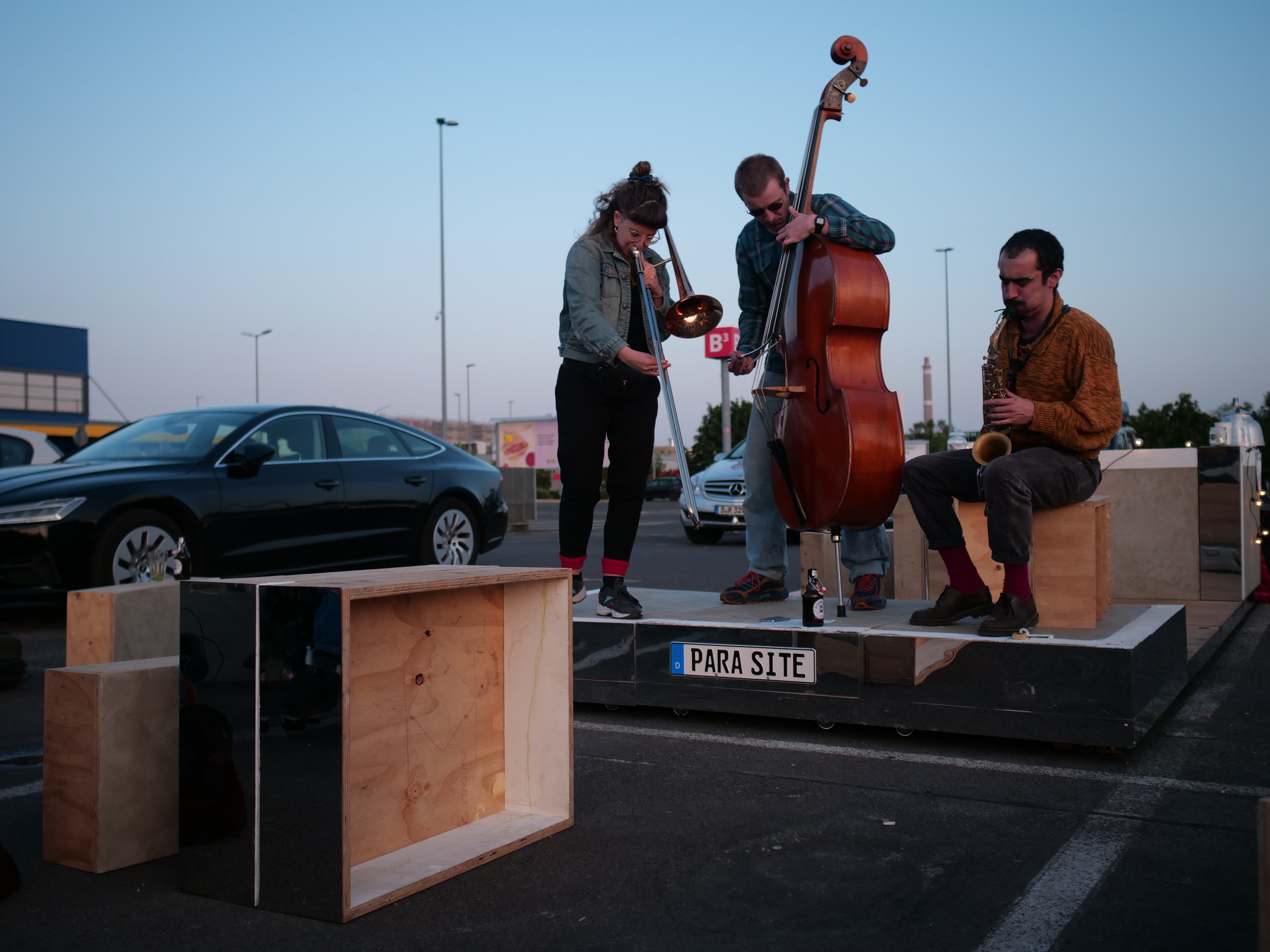 3 Personen musizieren, mit einem Kontrabass, einer Trompete und einem Saxophon auf der Plattform des Paraite Parkings. Diese steht auf einem Parkplatz vor einem Ikea.