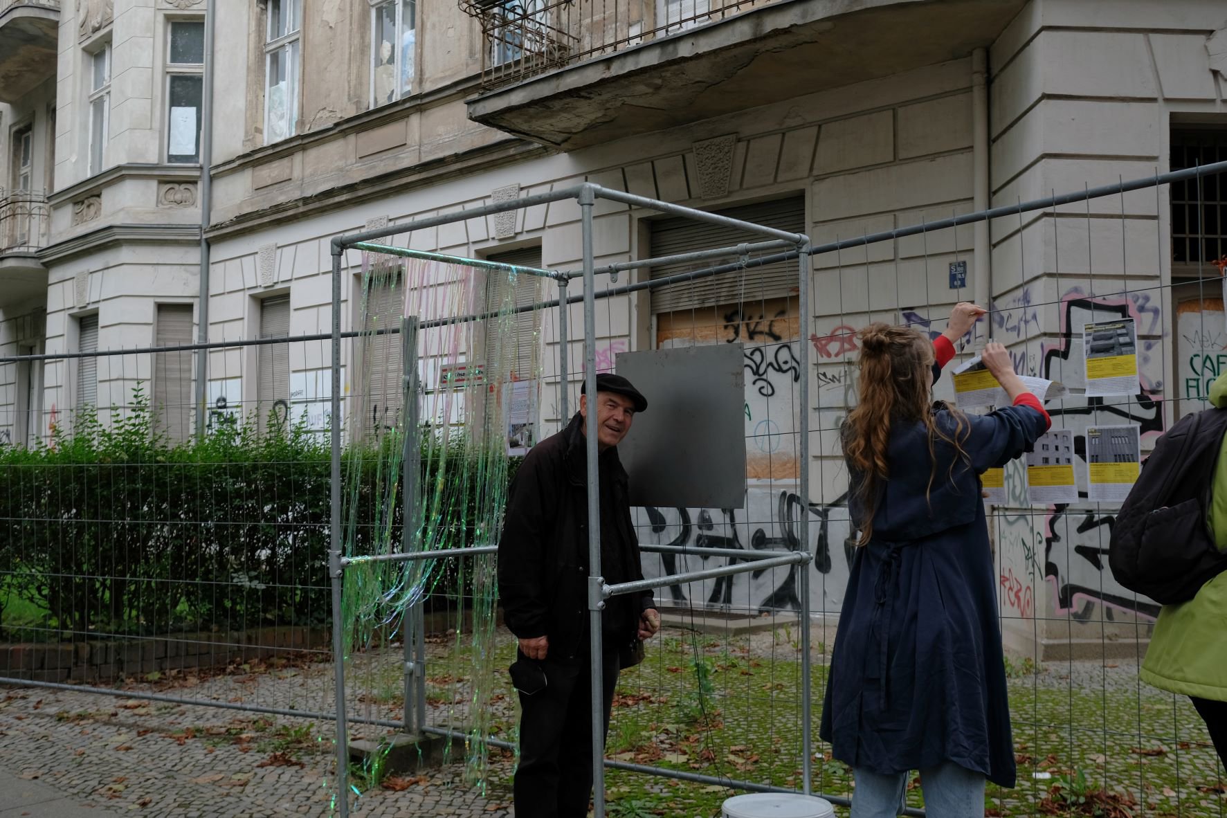 Vor einem leerstehenden Gründerzeithaus, das mit Graffitis besprüht ist, steht ein Bauzaun. An diesem steht ein älterer Mann in Mantel und Schiebermütze und guckt Richtung Kamera. Eine Frau hängt ein Plakat an den Zaun.