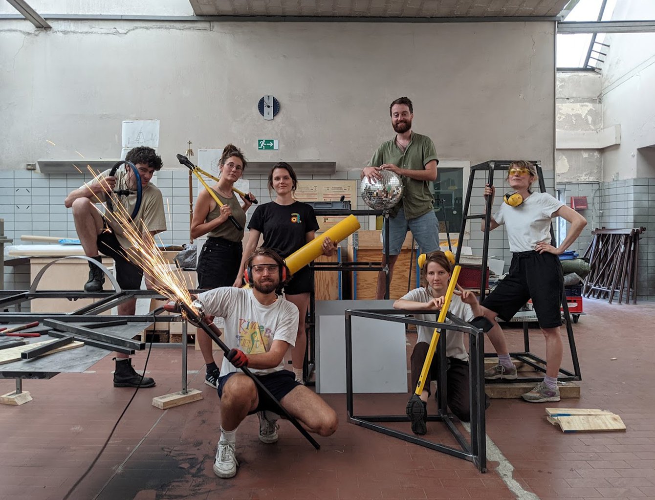 Auf dem Bild ist die 7-köpfige Karawane-Crew. Sie stehen in ihrer Werkstatt auf dem Dragoner Areal. Sie tragen Ohrschützer, Arbeitsbrillen und Werkzeuge sowie Holzbalken. Einer schmeißt gerade das Schweißgerät und versprüht Funken.