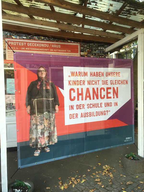 Auf einem großen Transpi am Gecekondu ist eine Person mit Hijab und Brille zu sehen. Neben ihr steht: Warum haben unsere Kinder nicht die gleichen Chancen in der Schule und in der Ausbildung? Darunter ist ihr Name, Zehra, zu lesen.