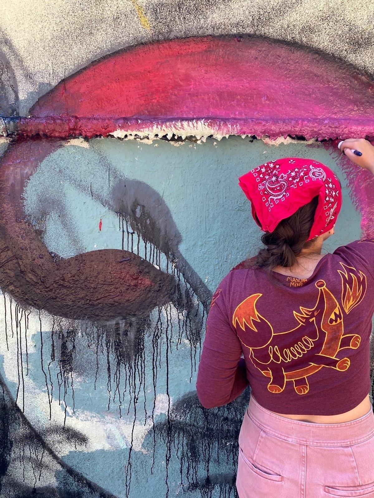 Ein ca 11-Jähriges Kind mit rosa Jeans, lila Hemd und Bandana im Haar malt einer braunen Hauswand ein buntes Graffiti. Man sieht das Kind von hinten.