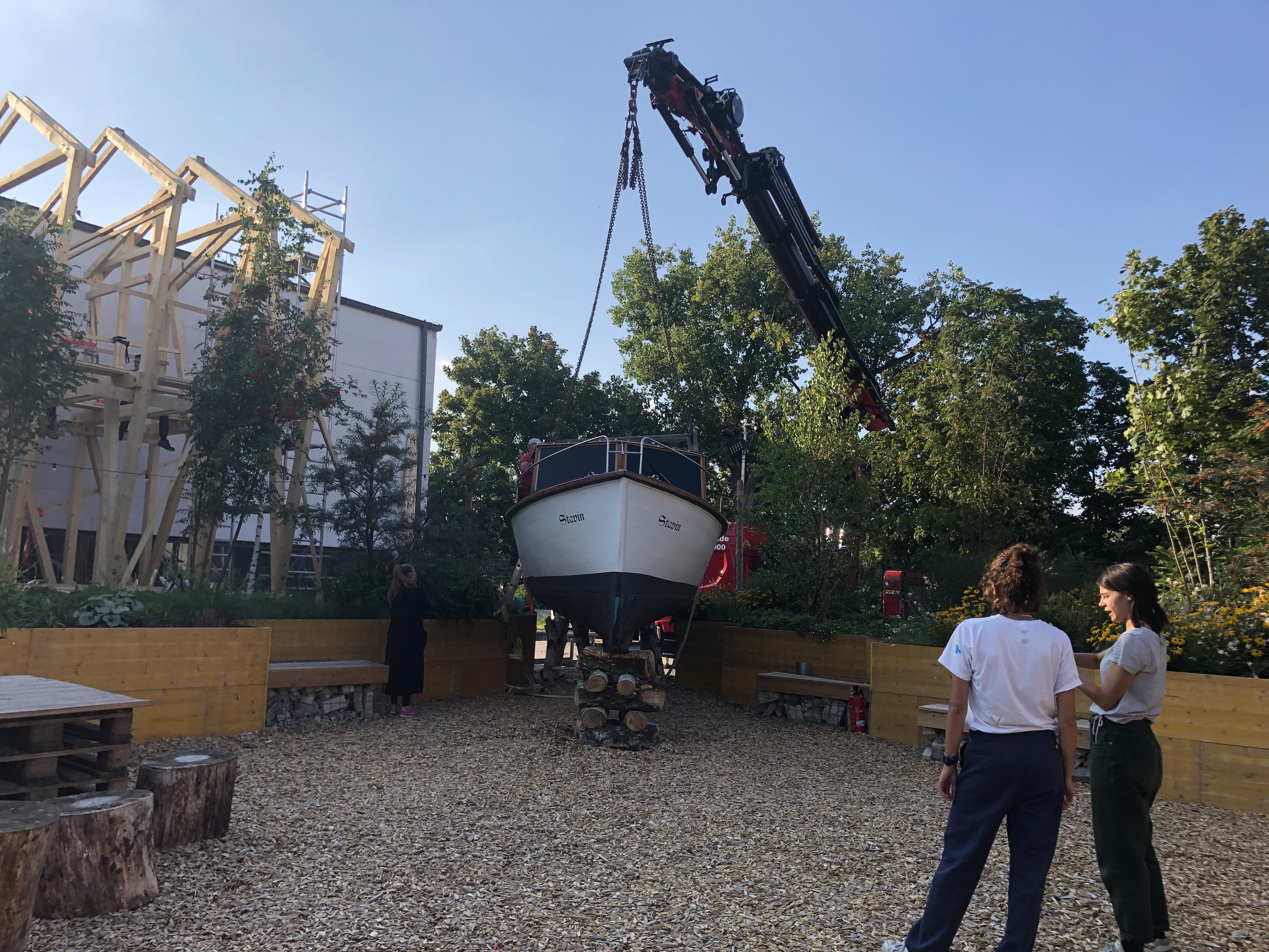 Zwei Menschen stehen mit dem Rücken zur Kamera rechts im Bild. Sie unterhalten sich und schauen wie das Boot mit dem Kran auf ein paar Holzstämme gestellt wird.