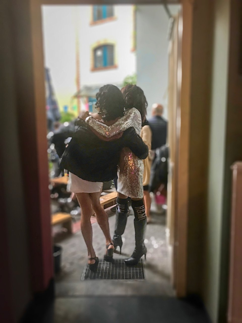 Zwei Personen mit langen schwarzen Haaren Arm in Arm von hinten.Sie stehen im Türrahmen vom Oya-Garten. Eine Person trägt ein kurzes Kleid und schwarze Jacke, die andere kniehohe Stilettostiefel und ein silbernes Paillettenkleid mit langen Ärmeln.