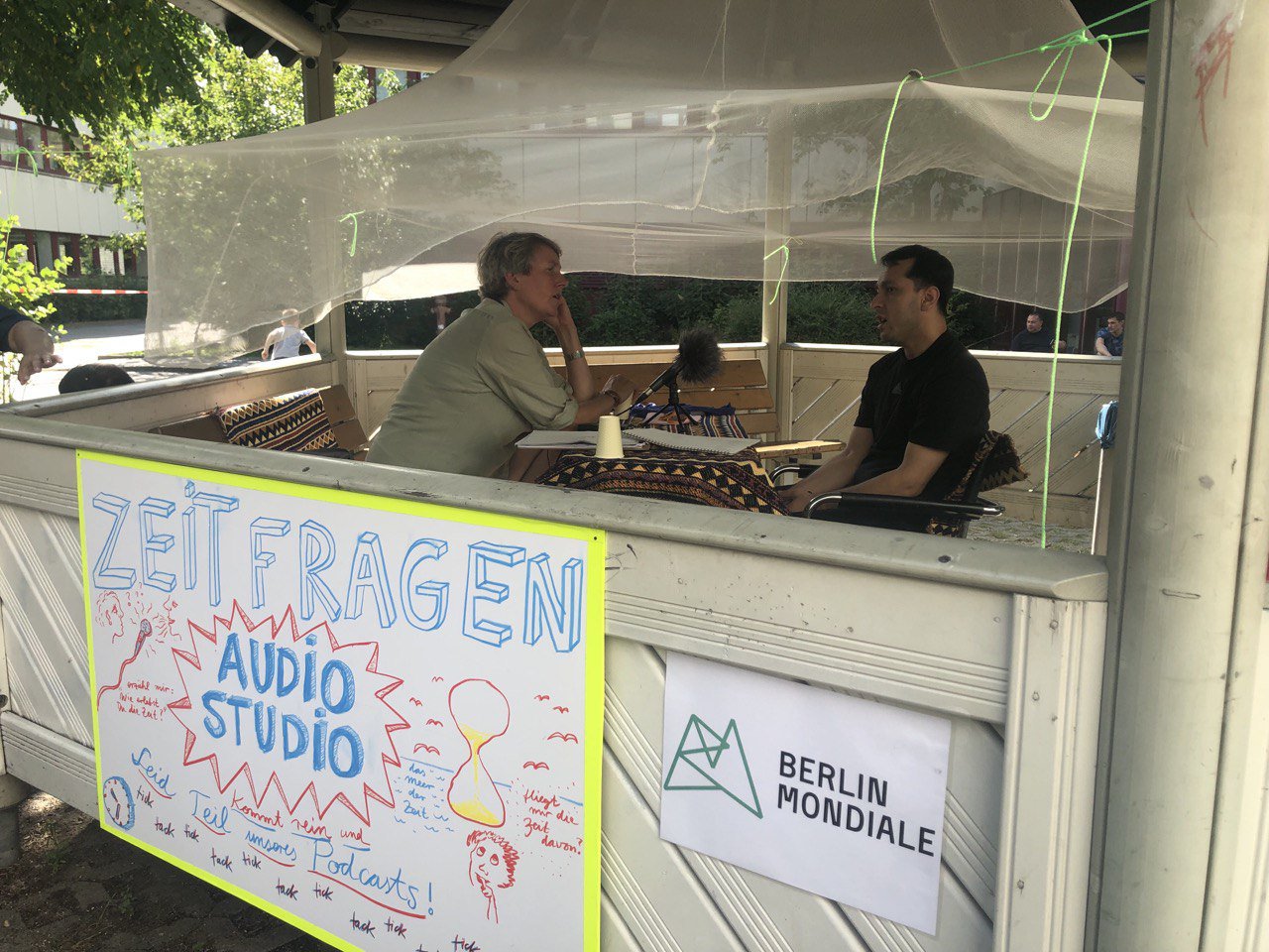 Ein Foto von zwei Personen, die den Podcast “Zeit fragen“ aufnehmen. Sie sitzen in einem Pavillon an einem Tisch, zwischen ihnen steht ein Mikrophon.