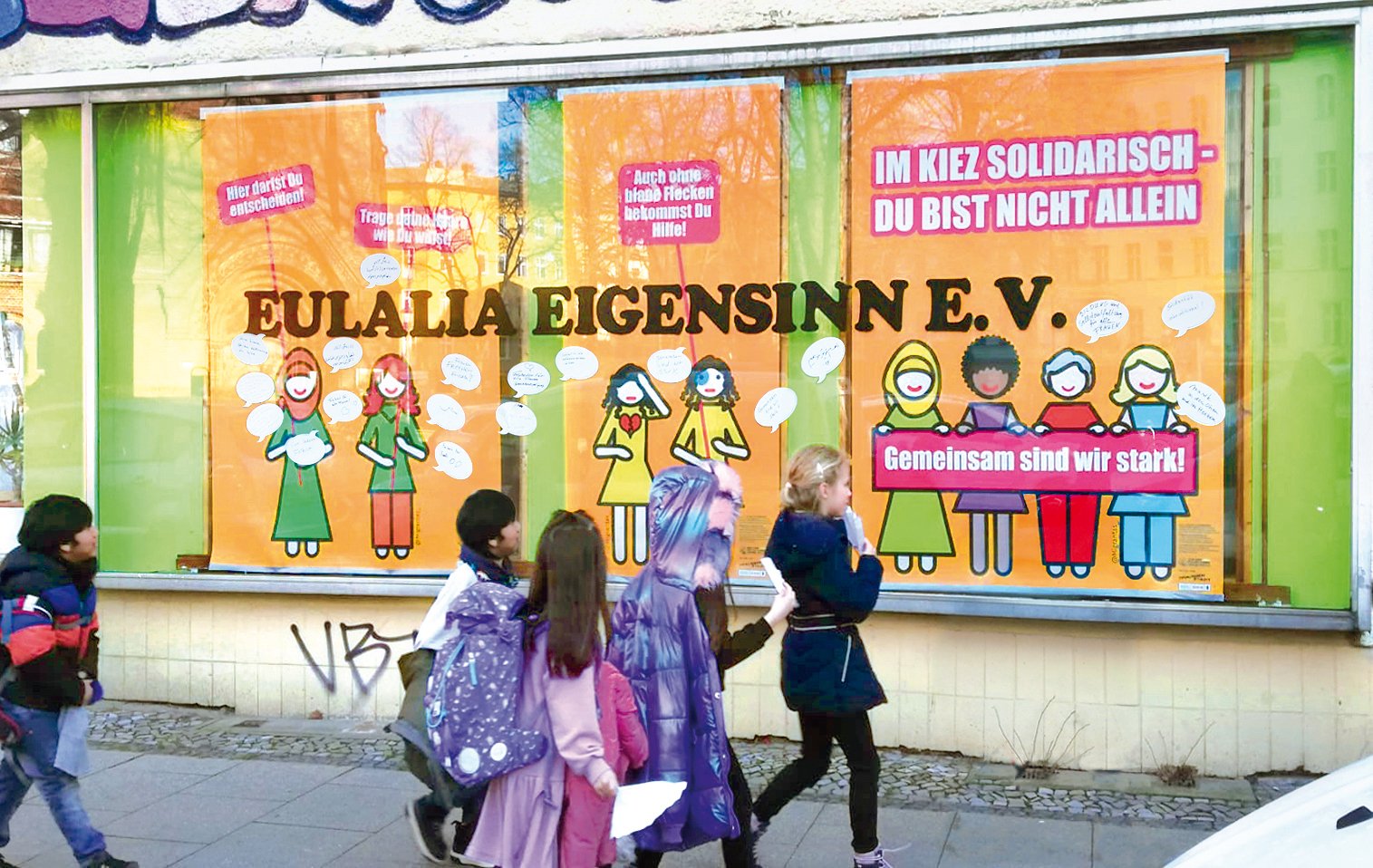 In einem Schaufenster sind drei große Plakate. Auf einem steht: Im Kiez solidarisch - du bist nicht allein. Darunter sind vier gezeichnete Personen mit unterschiedlichen Haaren, Kopfbedeckung und Hautfarbe. Diese halten ein Schild.
