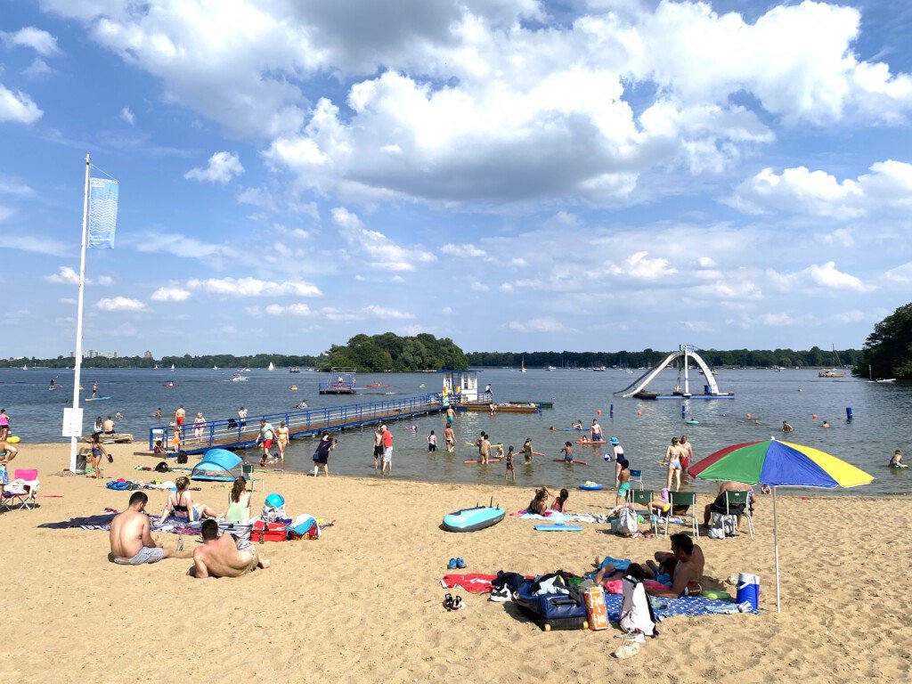 Das Bild zeigt das Strandbad Tegel. Im Vordergrund sitzen, stehen und liegen Menschen im Sand. Ein langer Steg mit einem Wasserwachthäuschen am Ende ragt ins Wasser. Im Hintergrund Segelboote.