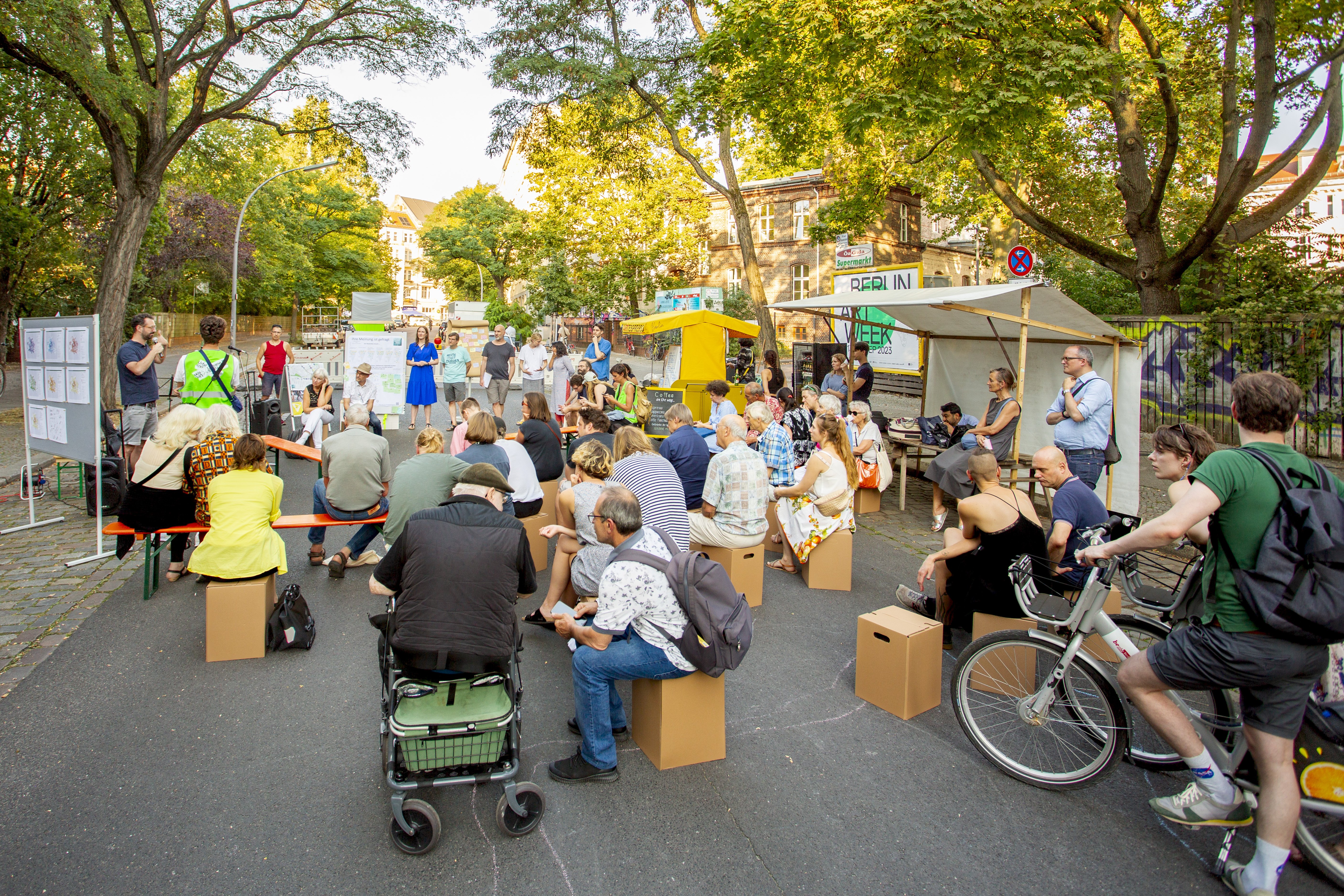 Auf einer Straße sind Bänke und Sitzhocker aufgebaut, auf denen Menschen sitzen. Sie schauen zu einer Person mit Mikro die vor einer aufgestellten Leinwand steht. Im Hintergrund sind weitere Leinwände und eine kleines Häuschen.