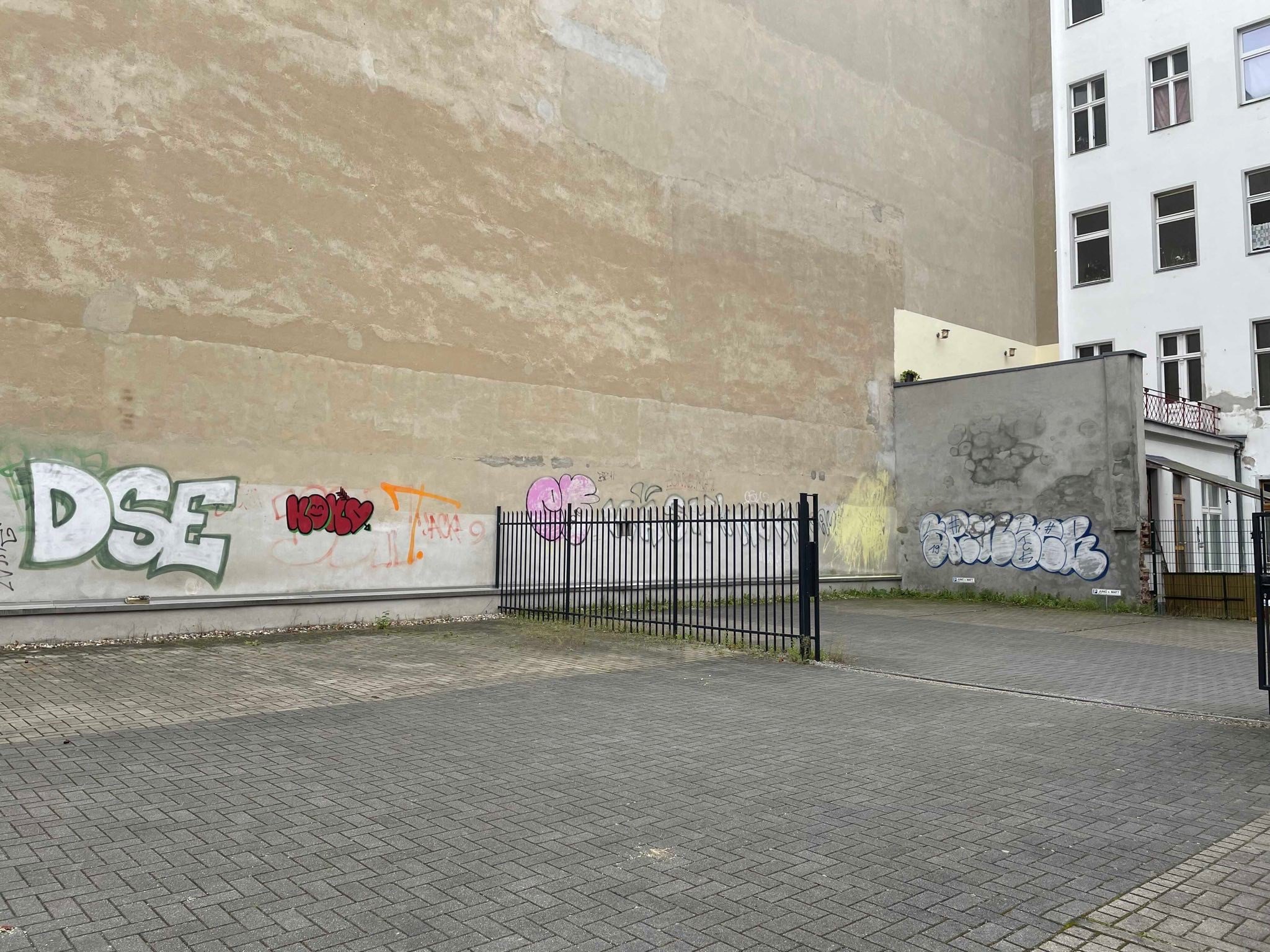 Ein Hinterhof mit einem schwarzen Zaun und einer großen hellbraunen fensterlosen Hauswand. Auf der Hauswand sind mehrere Graffitis. Der Hof bzw. Parkplatz ist gepflastert. Es ist kein grün zu sehen und der Hof ist leer.