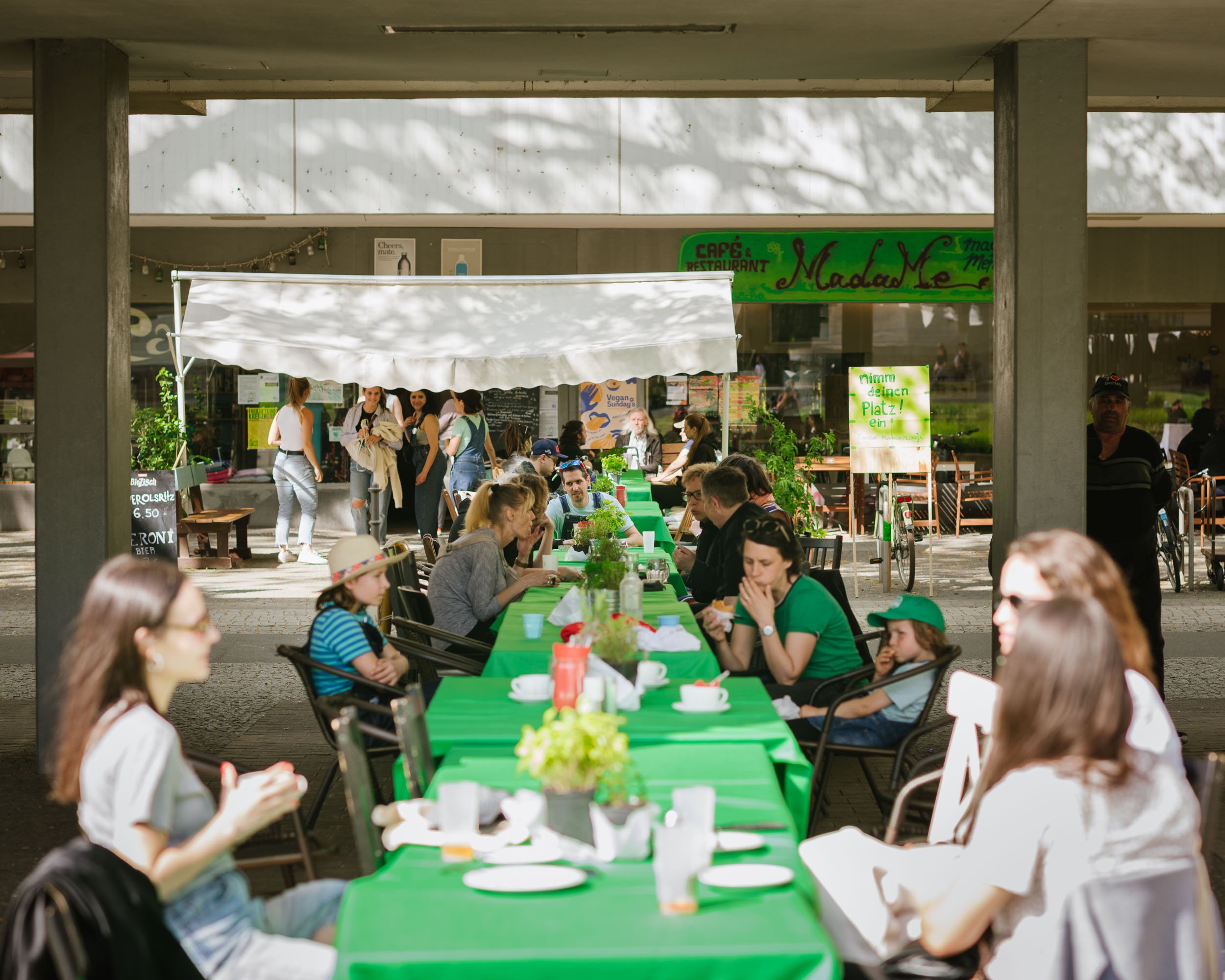Mehre Tische sind auf einem Gehweg längs aufgestellt. Sie haben alle grüne Tischdecken. An den Tischen sitzen verschiedene Menschen und quatschen und essen.