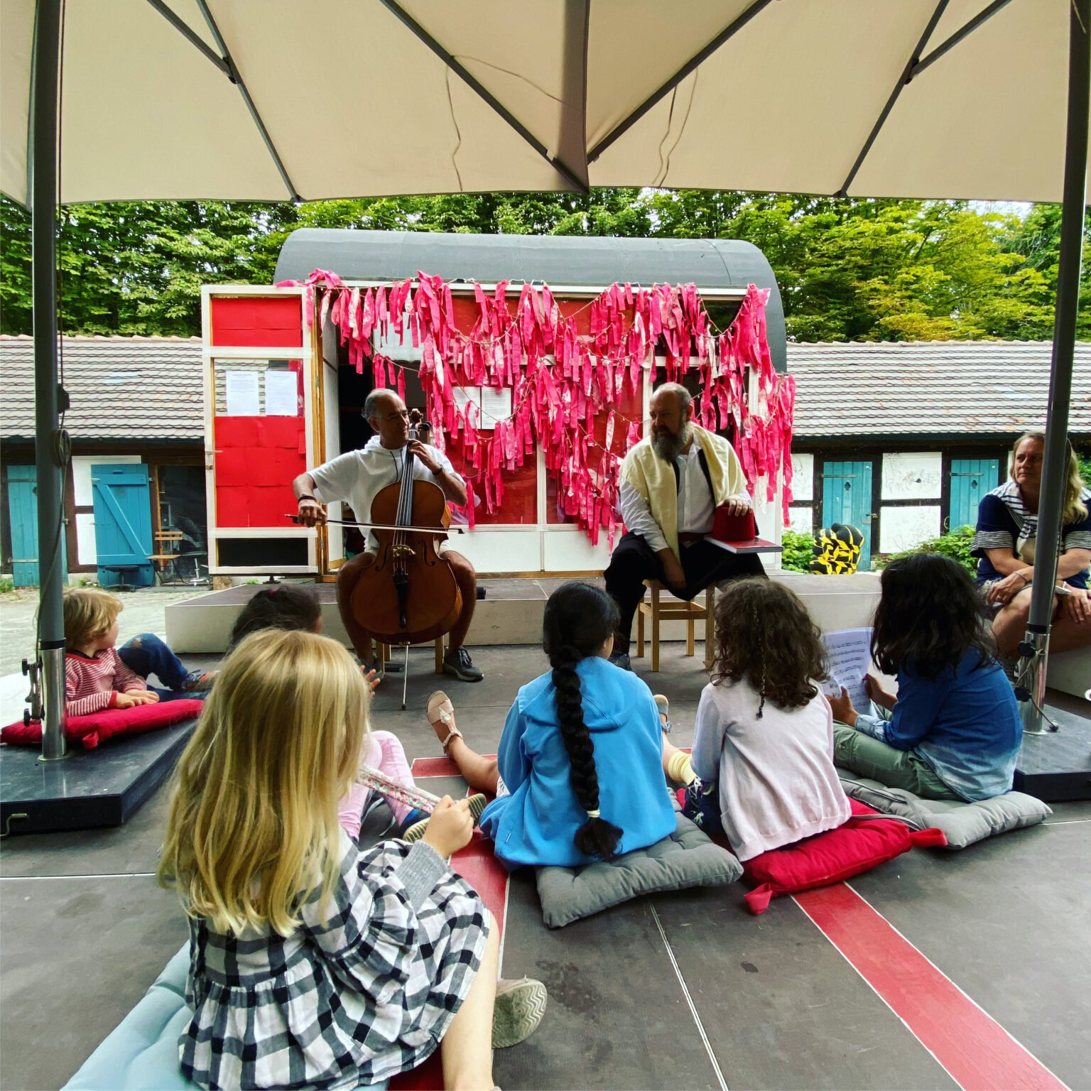 In der Spandauer Altstadt sitzen mehrere Kinder mit dem Rücken zur Kamera vor einer Freiluftbühne. Auf der Bühne sitzen zwei Männer auf Hockern. Dier eine spielt Cello, der andere hat ein Buch auf den Knien.
