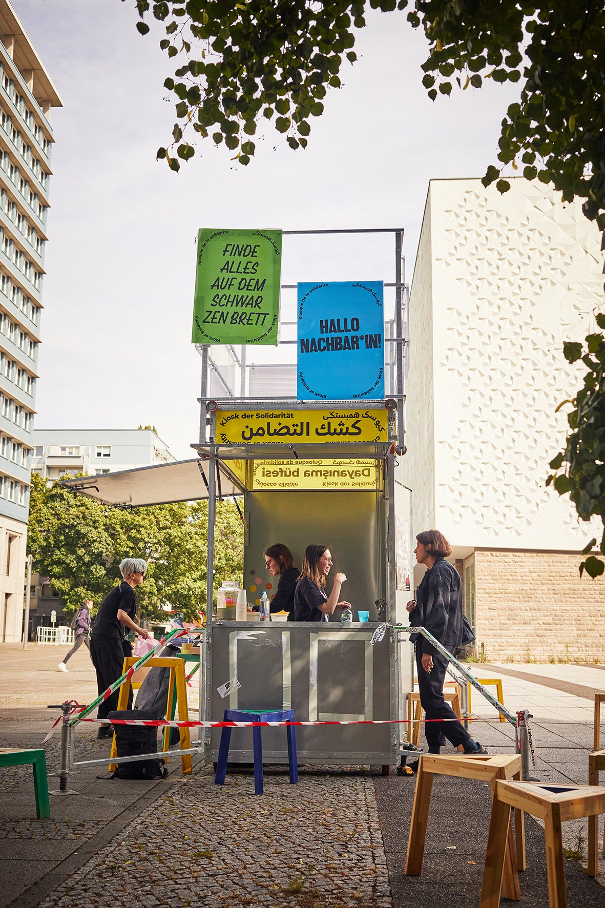 Das Baugerüst des Kiosk steht auf einem Bürgersteig. Davor kleine Hocker. Zwei Personen stehen im Kiosk. Zwei Personen stehen vor dem Kiosk. Auf dem oberen Teil des Kiosk sind Schilder, dort steht: "Hallo Nachbar*inne" und "Kiosk of Solidarity"