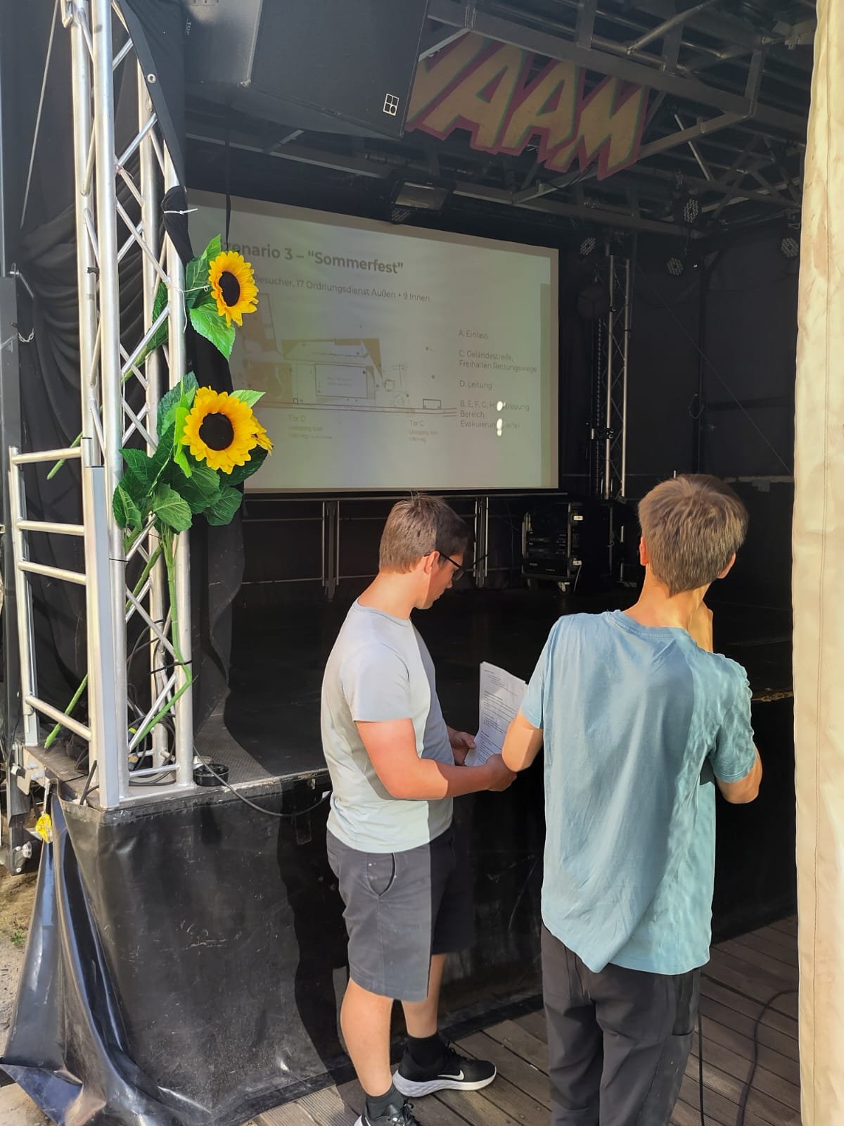Zwei Personen stehen vor einer Bühne. Auf der Bühne ist eine Präsentation gebeamed.