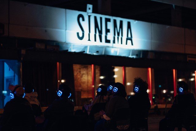 Viele Personen stehen vor dem Sinema im Haus der Statistik am Alexanderplatz. Sie tragen Kopfhörer. Es ist abends/nachts. Der Sinema-Schriftzug an der Hausfassade ist angeleuchtet, der Rest verschwommen im Dunklen.