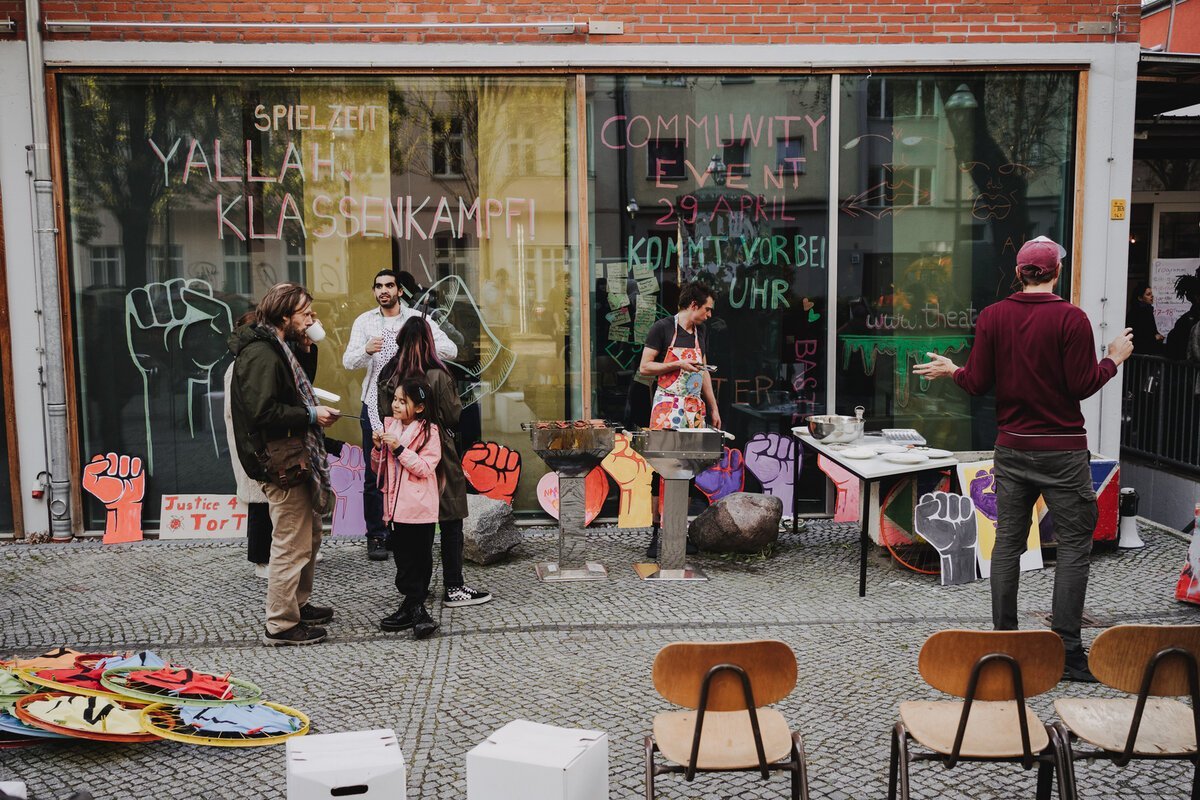 Auf einem Bürgersteig vor einem Haus mit breiter Glasfassade stehen 8 Personen, darunter ein Kind. Eine Person steht vor zwei Grills. Die anderen trinken Kaffee. Auf der Glasfassade steht:Community Event 29.April Kommt vorbei.Es gibt noch mehr Zeichnungen