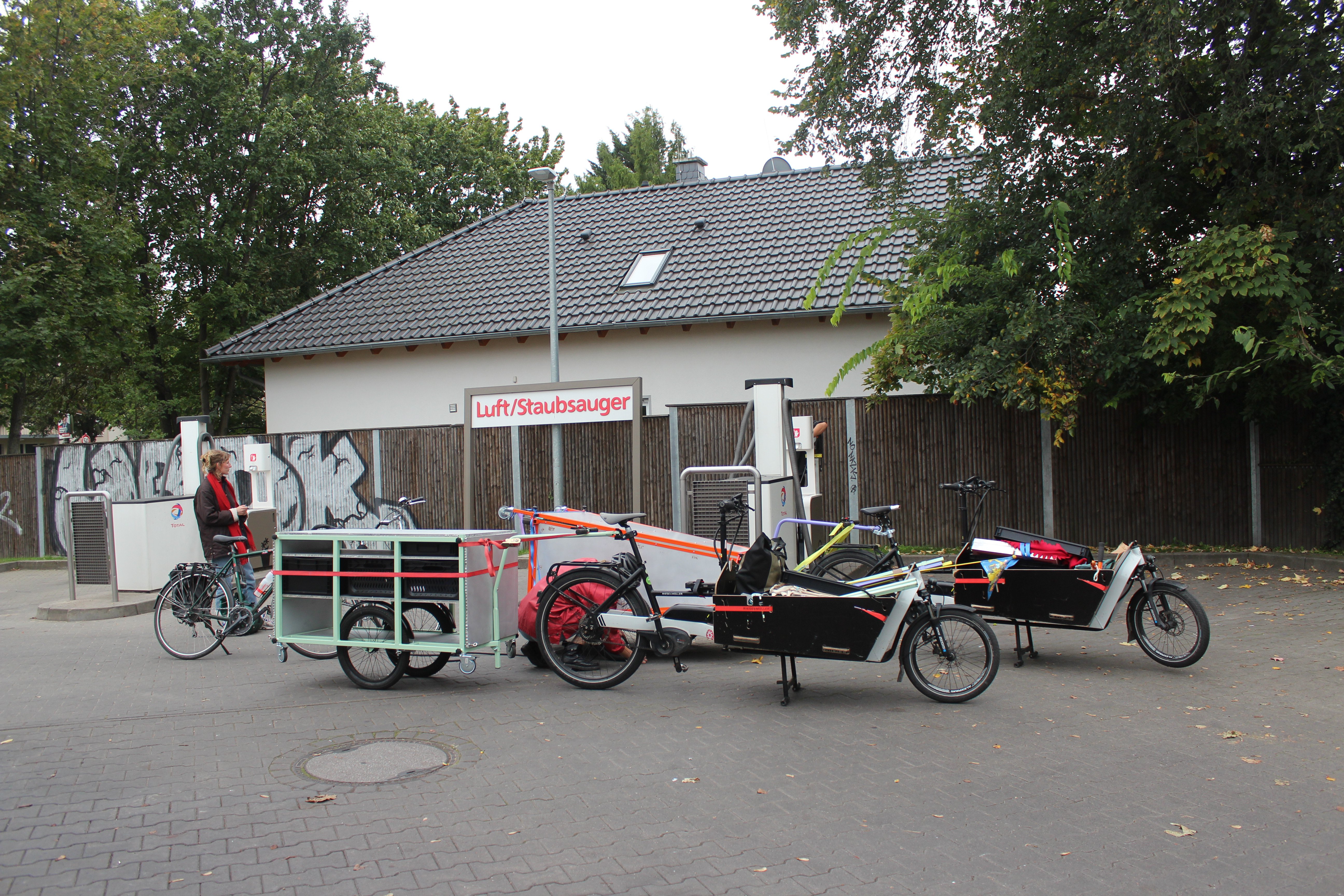 Auf einer Tankstelle vor der Luftpumpe stehen zwei Lastenräder mit Anhängern. Ein Anhänger ist die mobile Küche mit türkisem Holzgestell. Der andere Anhänger ist der Aufsteller aus organge-farbenen Holz. Dahinter steht eine Person mit Rad vor der Pumpe.