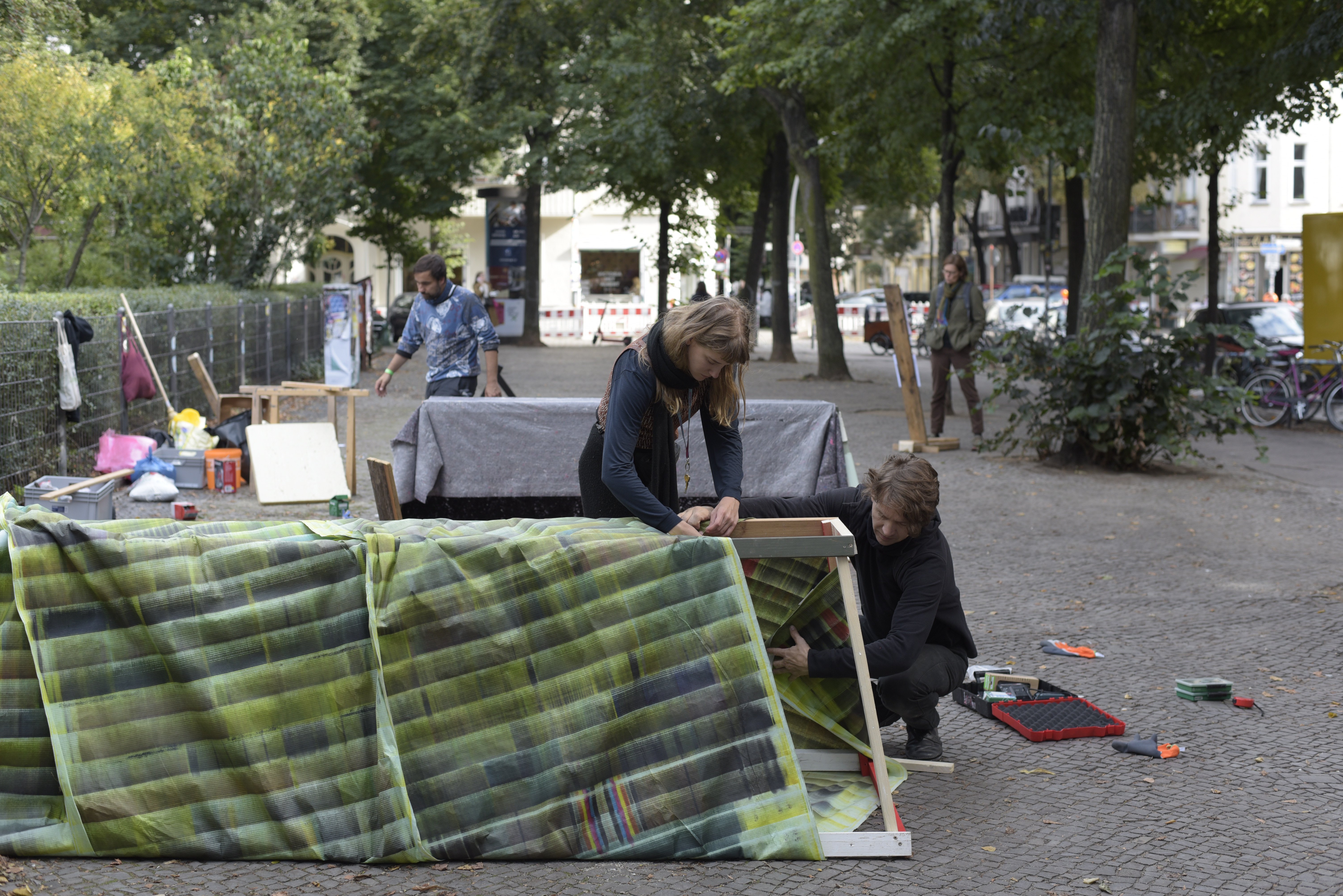 Auf einem gepflasterten Bürgersteig am Boxhagener Platz baut eine Person Skulpturen auf. Im Vordergrund verkleiden zwei Personen ein etwa 1,5 langes und etwa 1m hohes Holzgestell mit grünem Stoff.