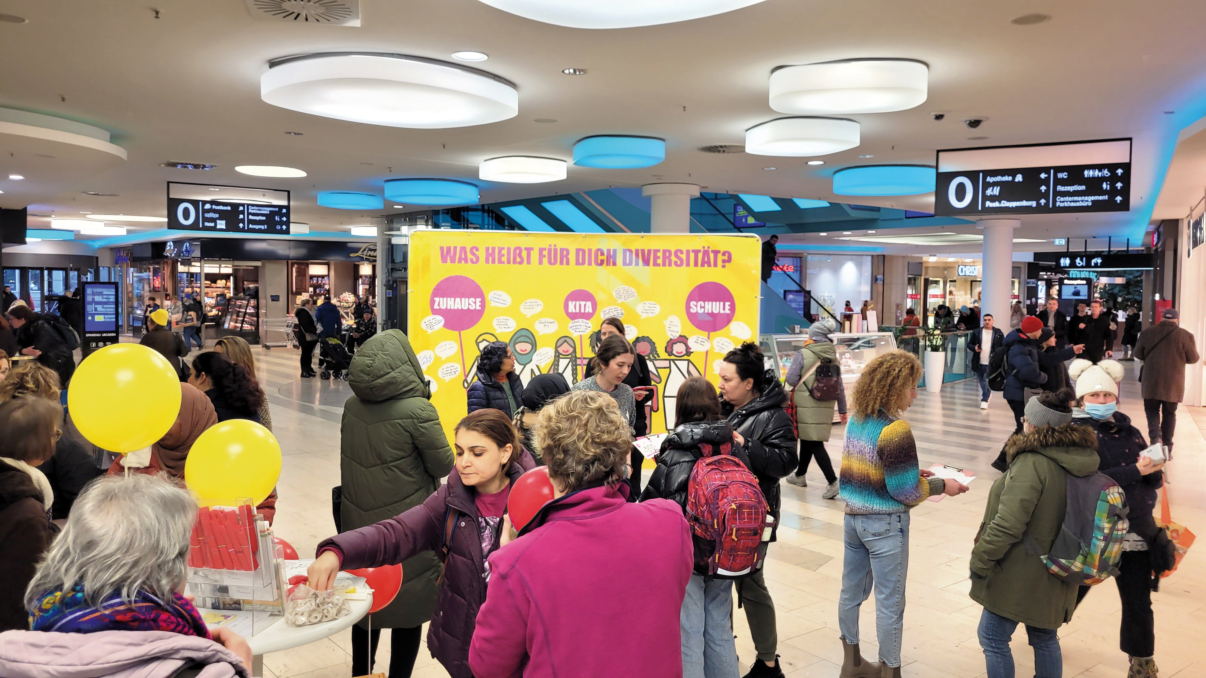 In einem Einkaufszentrum ist eine Plakatwand aufgestellt. Darauf steht: Was heißt für dich Diversität? Vor dem Plakat stehen verschiedene Menschen und reden.