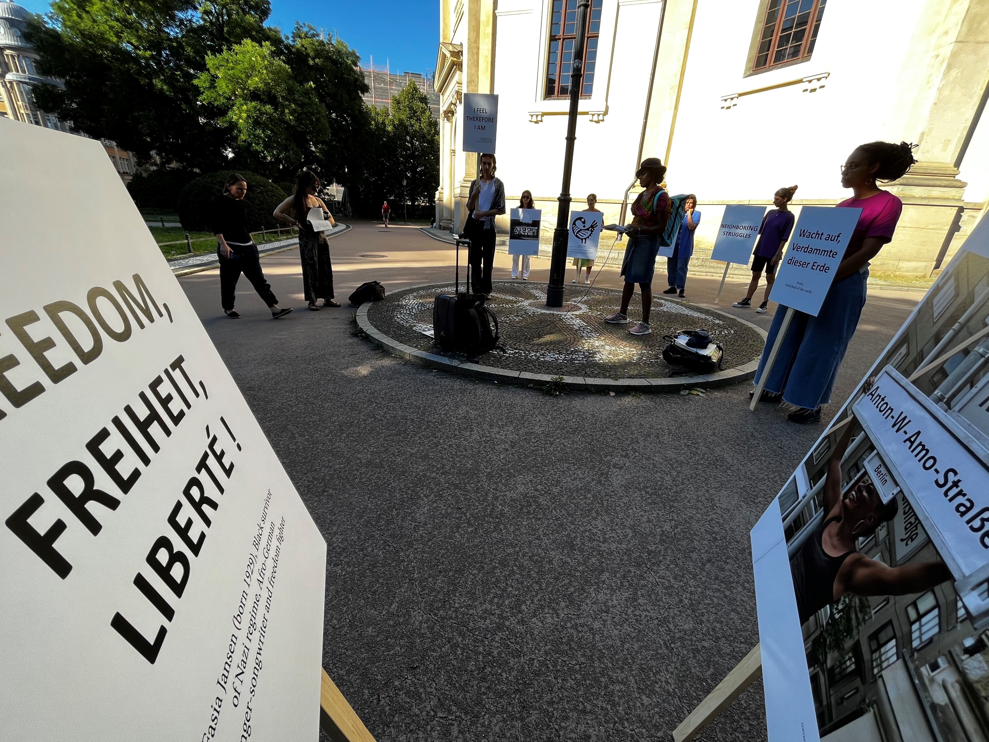9 Menschen stehen auf einem Platz. Sie halten Schilder mit Sätzen wie "Wacht auf, Verdammte dieser Erde" und "I feel therefore I am". Im Vordergrund des Fotos stehen zwei große Tafeln mit den Beschriftungen "Freedom,Freiheit, Liberté!" und Fotos.