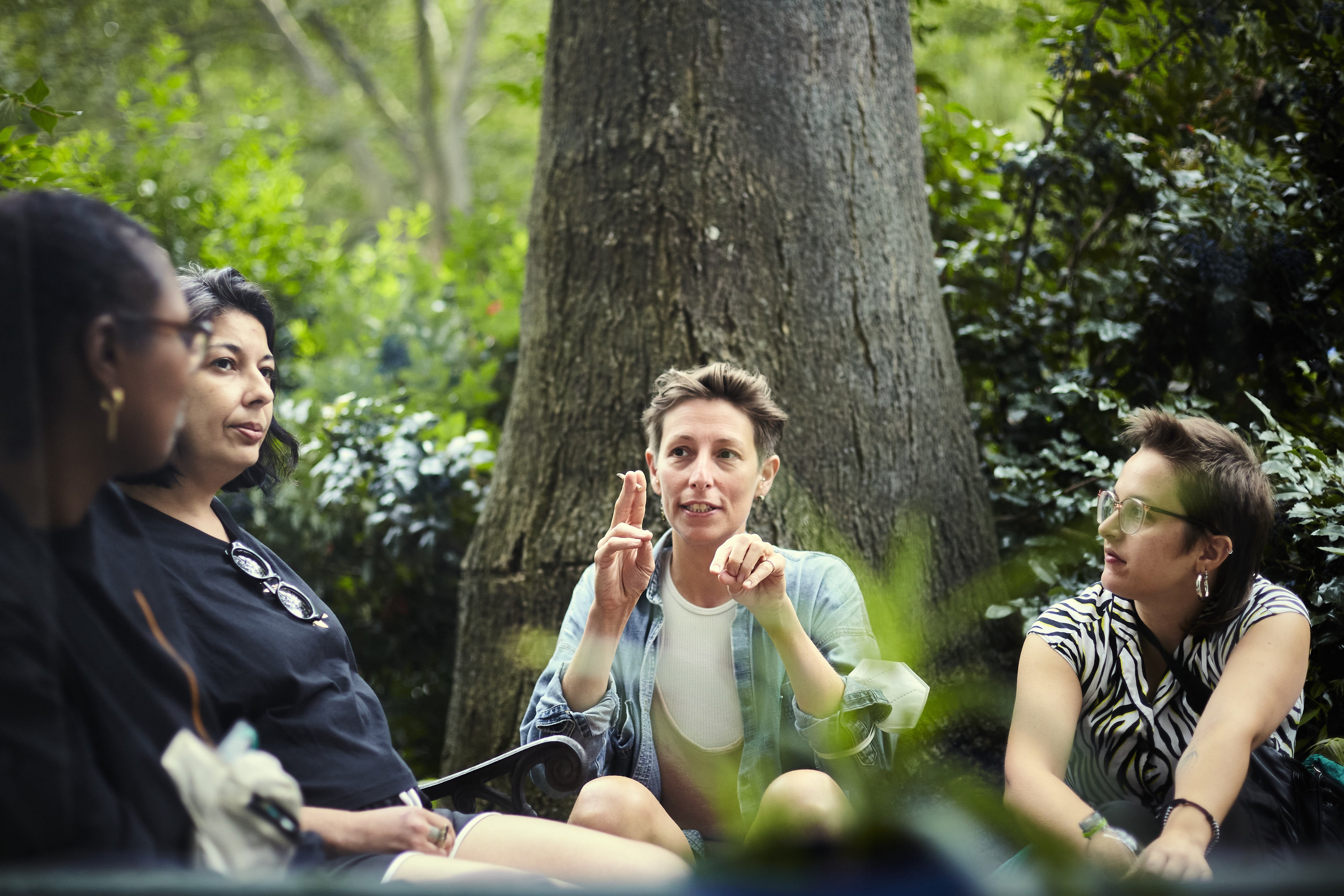 2.	Das Bild ist draußen aufgenommen und zeigt in Nahaufnahme vier Personen, die unter einem Baum sitzen. Eine Person mit kurzen hellen Haaren ist am gestikulieren, die anderen scheinen der Person zuzuhören.