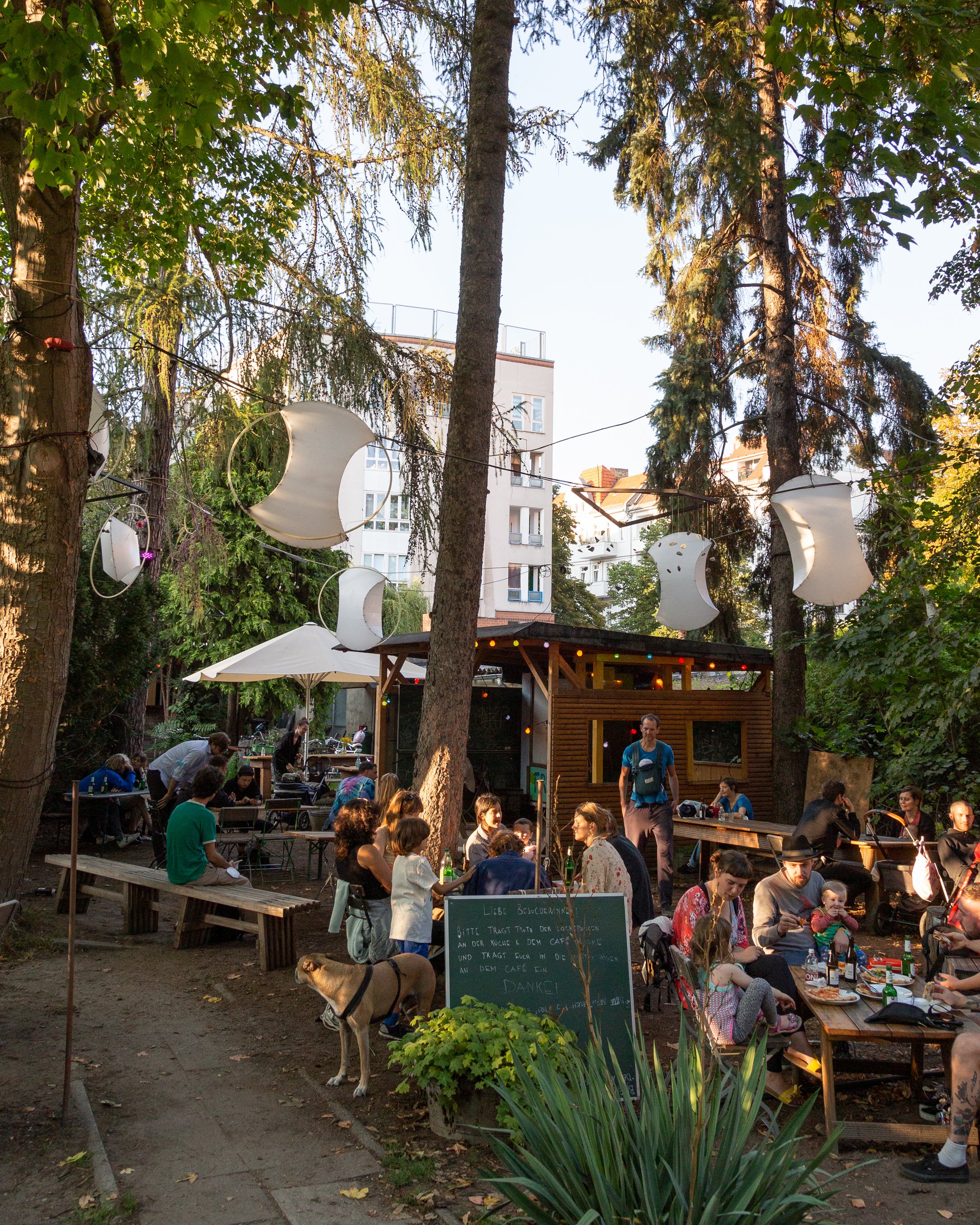 Das Bild zeigt die Prinzessinnengärten in Neukölln im Sommer. In der Mitte ist der Kiosk. Zwischen den Bäumen sitzen Erwachsene und Kinder in Kleingruppen an Tischen. Darüber hängen weiße Lampions.