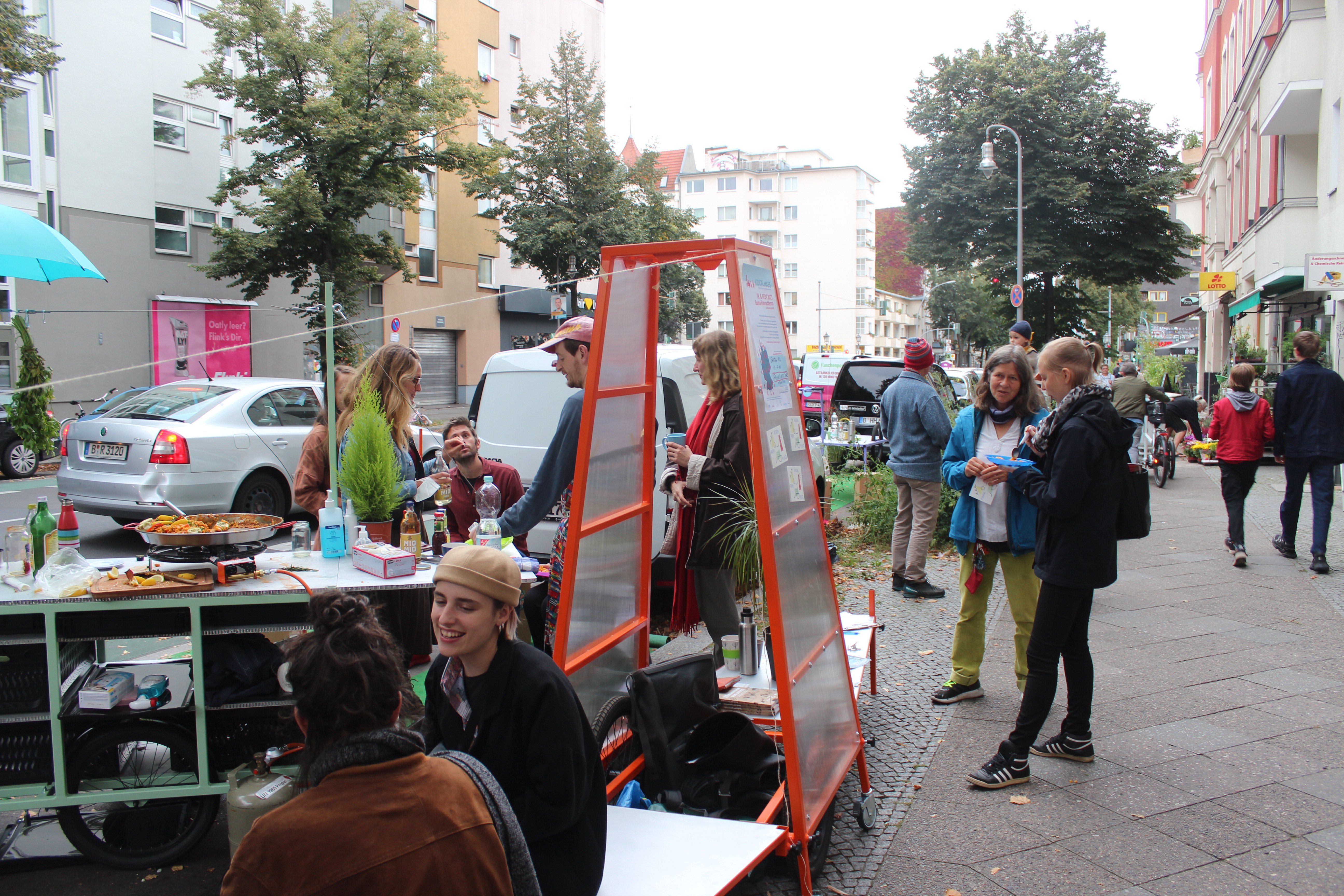 Die gleiche Szene aus dem vorigen Bild aus einer anderen Perspektive. Auf dem Fußweg neben dem Parkplatz steht ein Rad-Anhänger, der als Plakat-Aufsteller fungiert. Das Gestell ist aus Holz und orangefarben, die Wände transparent.