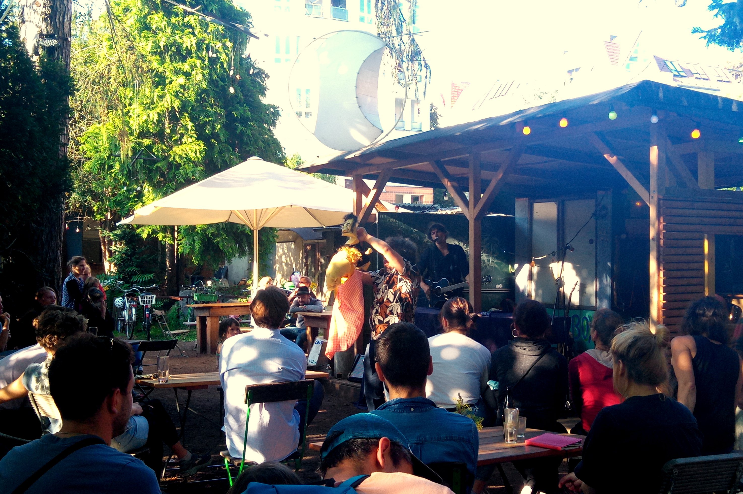 Auf der Open-Air-Bühne in den Prinzessinnengärten steht bei Sonnenschein eine Person mit Gitarre und singt. Vor der Bühne steht eine Person, die zwei Handpuppen bzw. eine Maske bewegt. Im Publikum sitzen ca. 20 Leute.