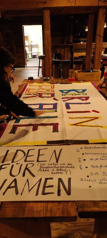 Auf einem Tisch, im inneren eines Raums, liegen zwei große Plakate. Auf einem steht "Berlin für Alle" und auf dem anderen "Ideen für Namen" und weitere Stichpunkte. Links malt eine Person an dem "Berlin für alle" Plakat weiter