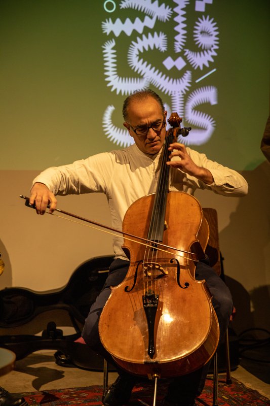 Ein Mann sitzt auf einer Bühne. Hinter ihm ist eine Illustation auf grünen Hintergrund gebeamt. Er spielt ein Cello.