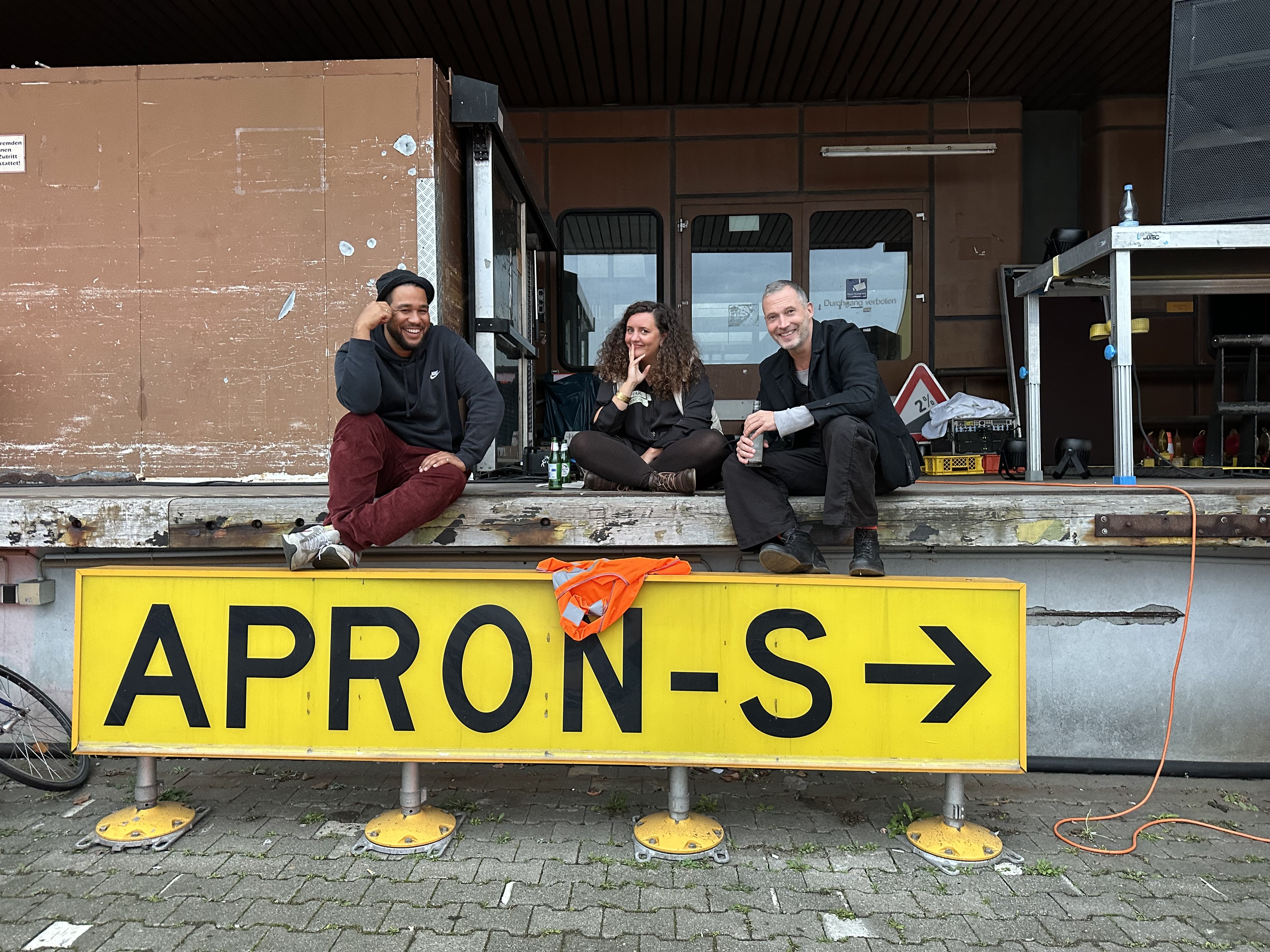 Drei Mitarbeitende des Berliner Projektfonds Urbane Praxis sitzen auf der Ladefläche der TXL Fläche. Unter ihren Füßen ist ein Schild auf dem Apron-S und ein Pfeil nach rechts steht.