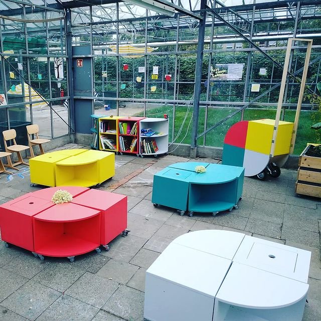 In einem Gewächshaus auf dem Gelände des Freiraumlabors stehen 4 Holzkisten zum Sitzen in den Farben rot, grau, gelb und türkis. Links im Hintergründe sind drei Schul-Stühle. Im Hintergrund mittig ist ein niedriges Bücherregal im Stile der Holzkisten.