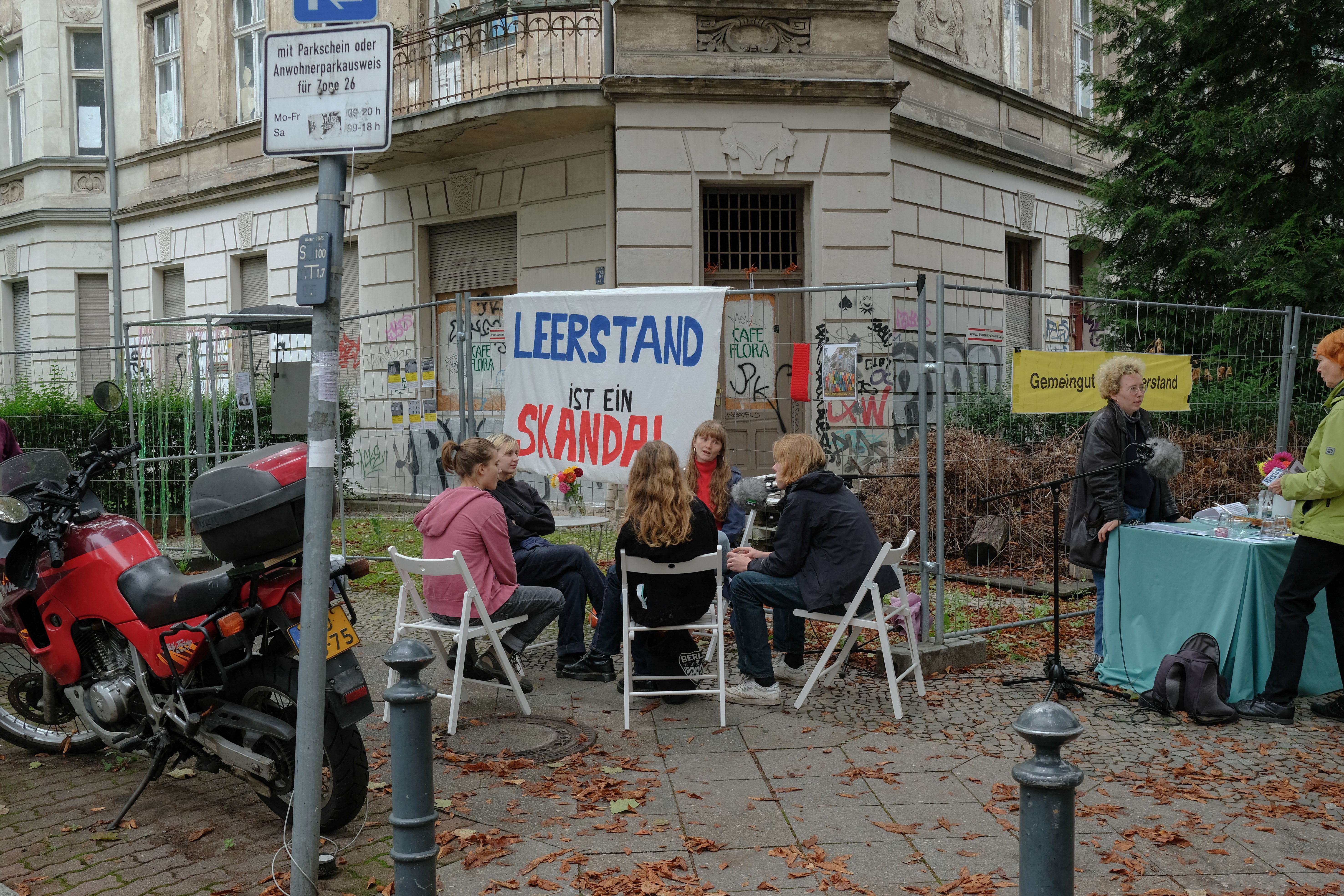 Vor einem leerstehenden Haus in Schöneberg sind Bauzäune aufgestellt. Am Zaun hängt ein großes Transparant mit Aufschrift: Leerstand ist ein Skandal. Davor unterhalten sich 5 Person im Stuhlkreis. Rechts am Zaun steht "Gemeingut statt Leerstand".