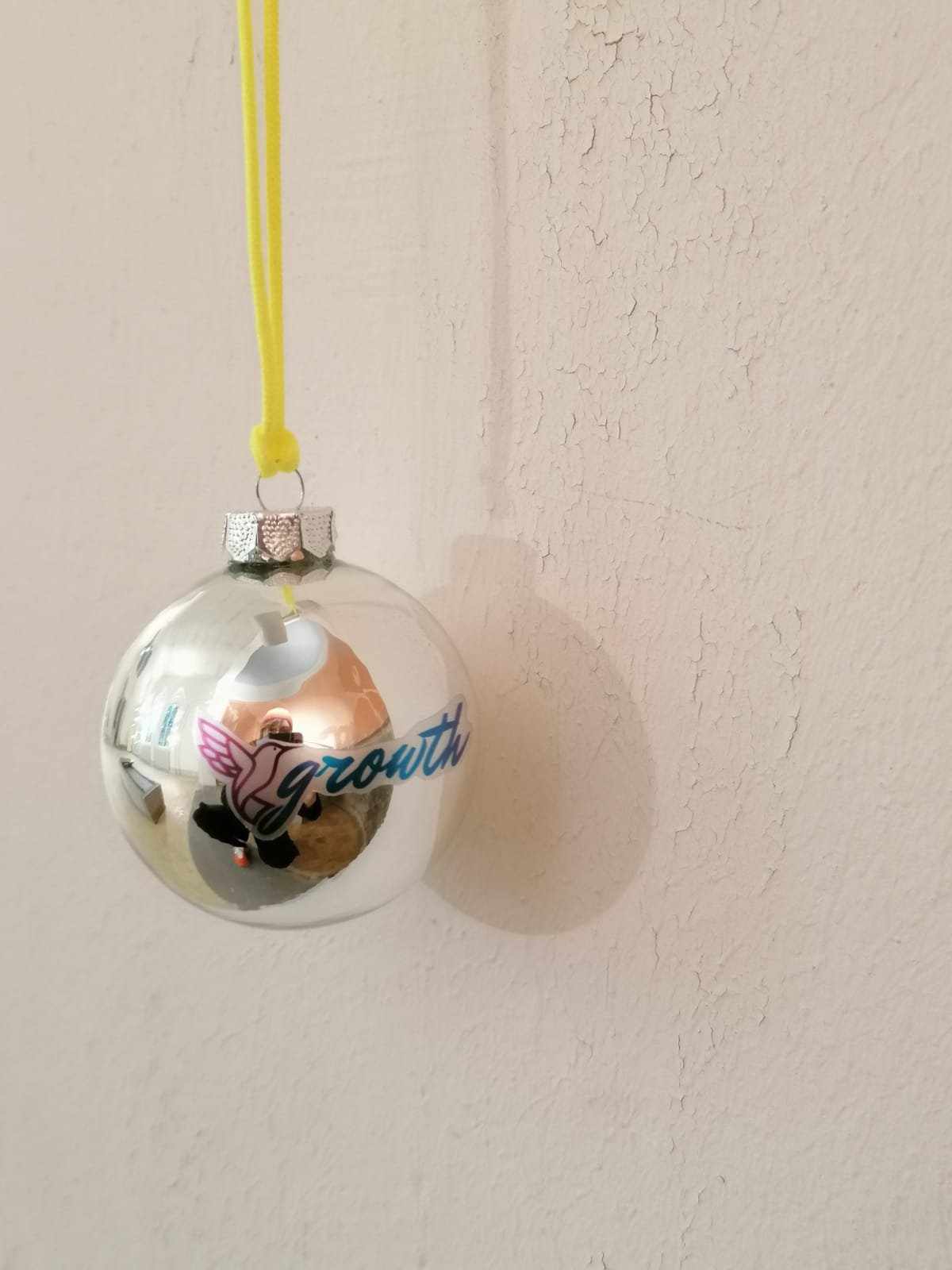 Nah-Aufhnahme: Eine silberne Weihnachtskugeln mit dem Aufdruck "growth" hängt vor einer weißen Wand.