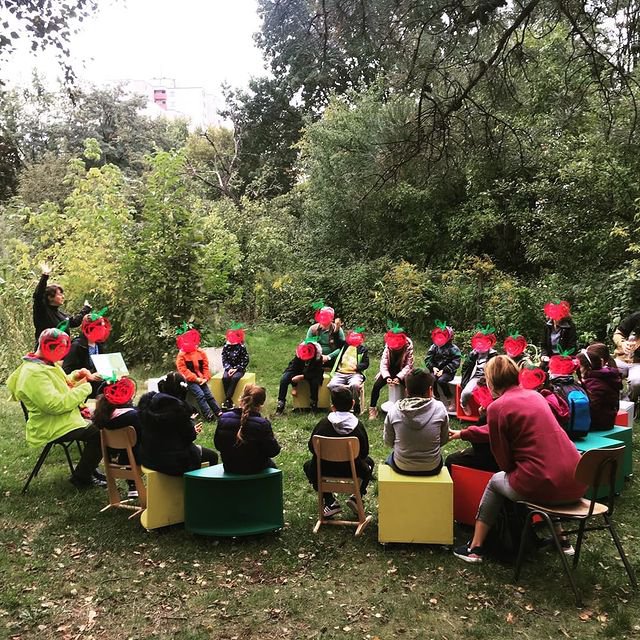 Eine Gruppe von 15 Kindern und Erwachsenen sitzen in einem Garten im Kreis. Die Gesichter sind durch Sticker in Apfelform unkenntlich gemacht. Eine Person zeigt den Kindern Abbildungen in einem Buch.