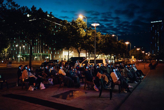 Auf dem Weg vor dem Sinema sitzen bei Dämmerung etwa 50 Personen in Reihen auf Plastikstühlen. Die Personen tragen Kopfhörer, deren Umrandung beleuchtet ist und schauen Richtung Fassade des Sinemas.