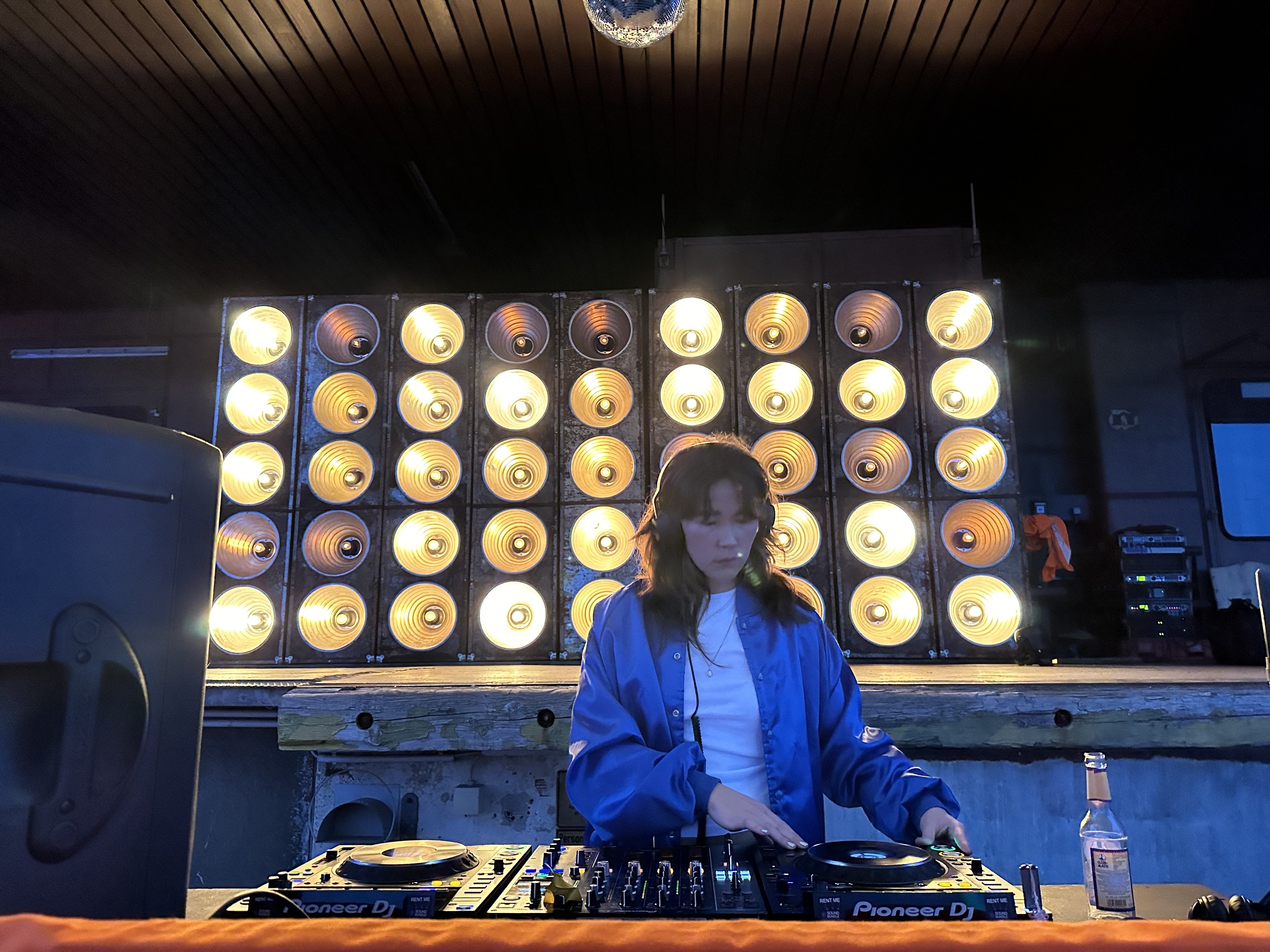 Eine Frau steht am einem DJ Pult und bedient dieses. Im Hintergrund ist auf einer Plattform eine Wand aus Scheinwerfern.