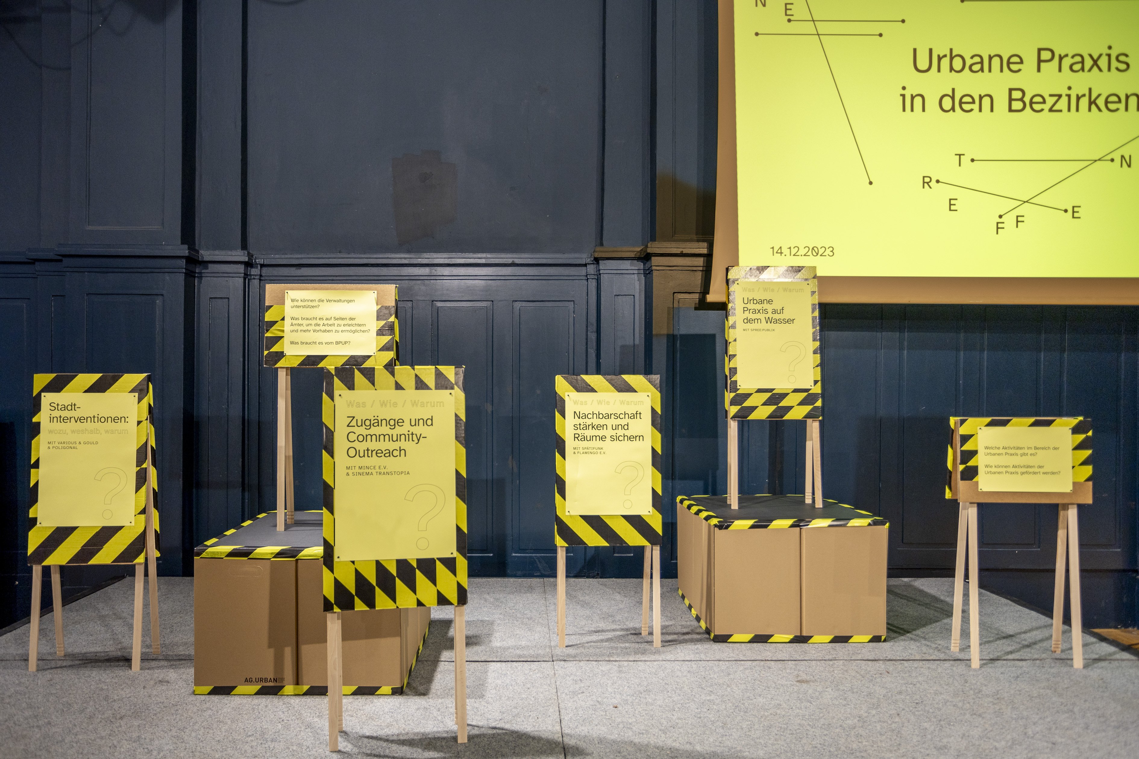 Es stehen 6 Schilder auf Holzbeinen auf einem Podest. Die Schilder haben gelb-schwarzes Flatterband aufgeklebt, darüber sind gelbe Plakate geklebt auf denen Themen stehen. Beispielsweise: "Zugänge und Community-Outreach" oder "Stadtinterventionen".