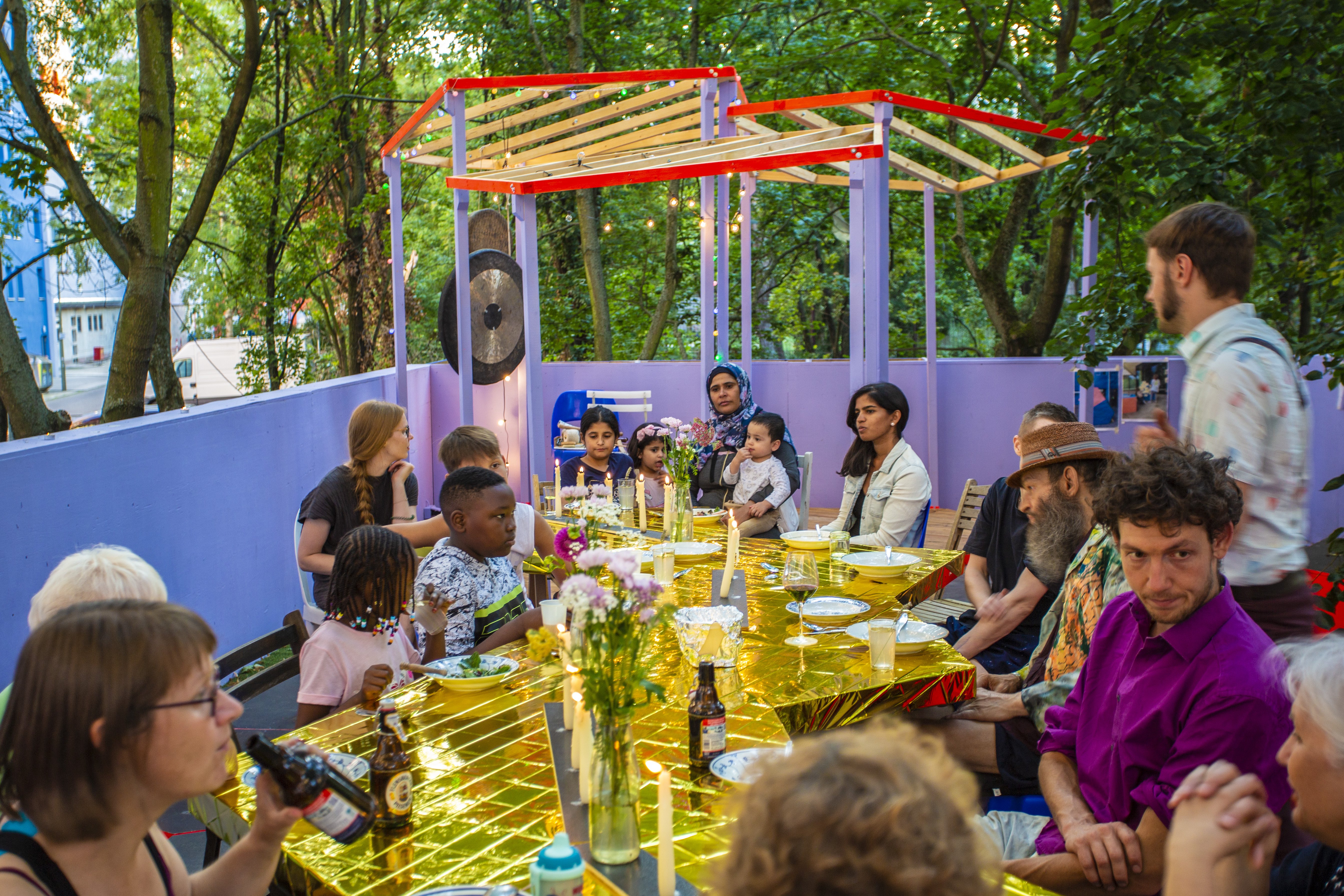 Das Foto zeigt einen in gold gedeckten Essenstisch mit Kindern und Erwachsenen aus der Nachbarschaft. Die Fläche um den Tisch ist mit halbhohen lila Holz-Mauern begrenzt.