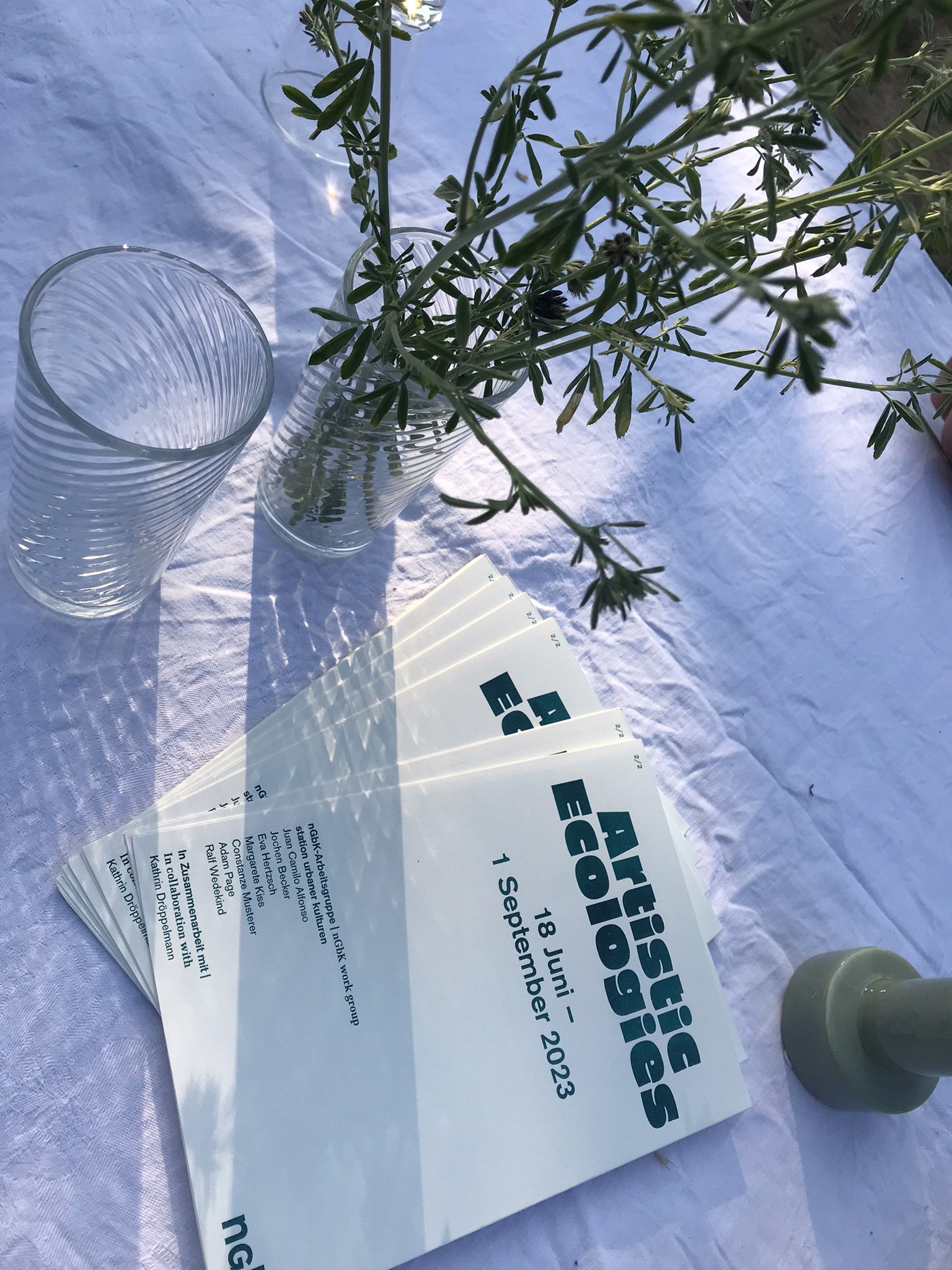 Ein Tisch mit Tischdeck. Auf ihm steht ein Glas mit Blumenstrauß, ein Glas und ein Heft auf dem fett "Artistic Ecologies" steht