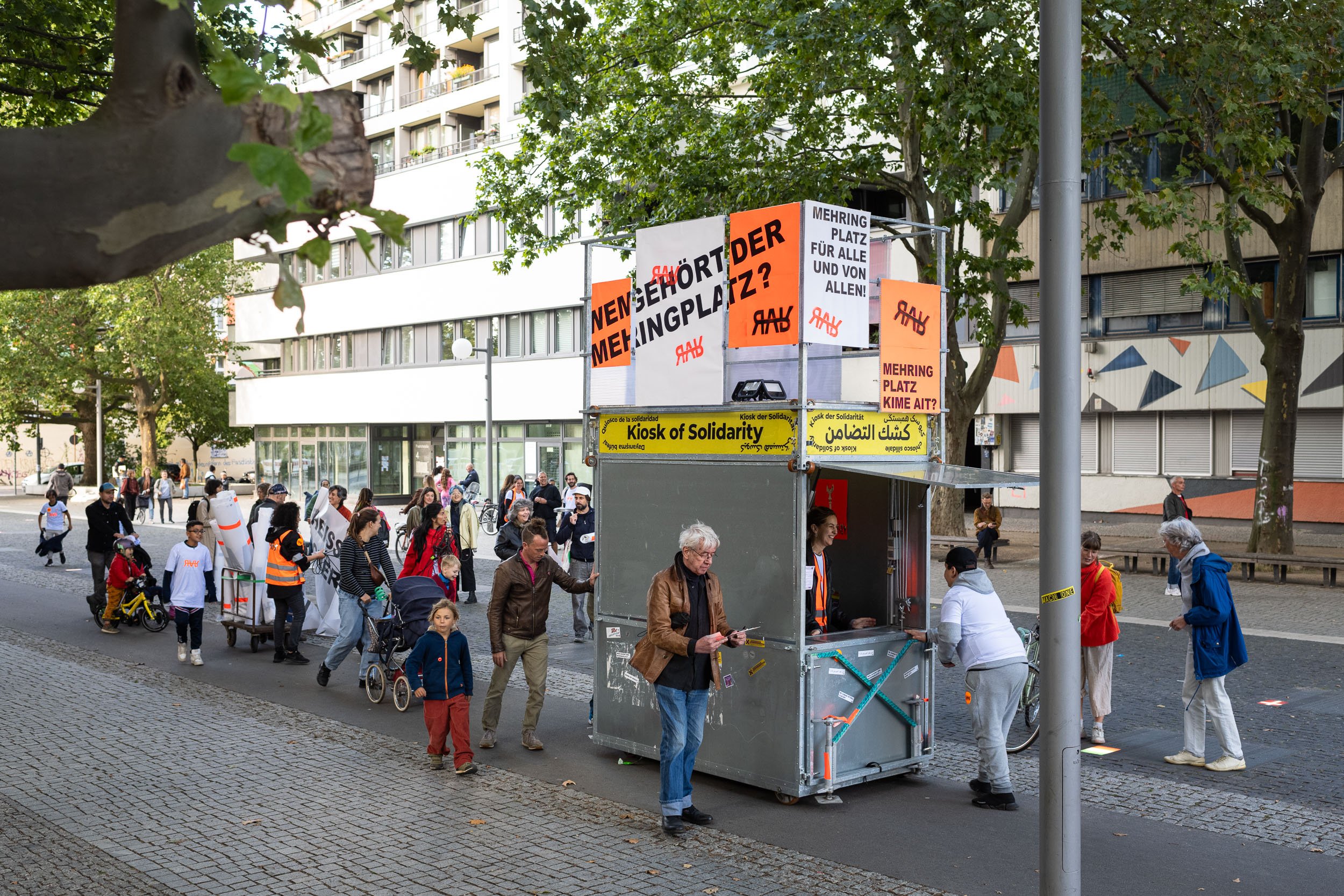 Das Gerüst des Kiosk of Solidarity steht auf einem Bürgersteig. Darum sind Menschen versammelt, die miteinander reden, den Kiosk anschauen oder mit der Person im Kiosk reden.