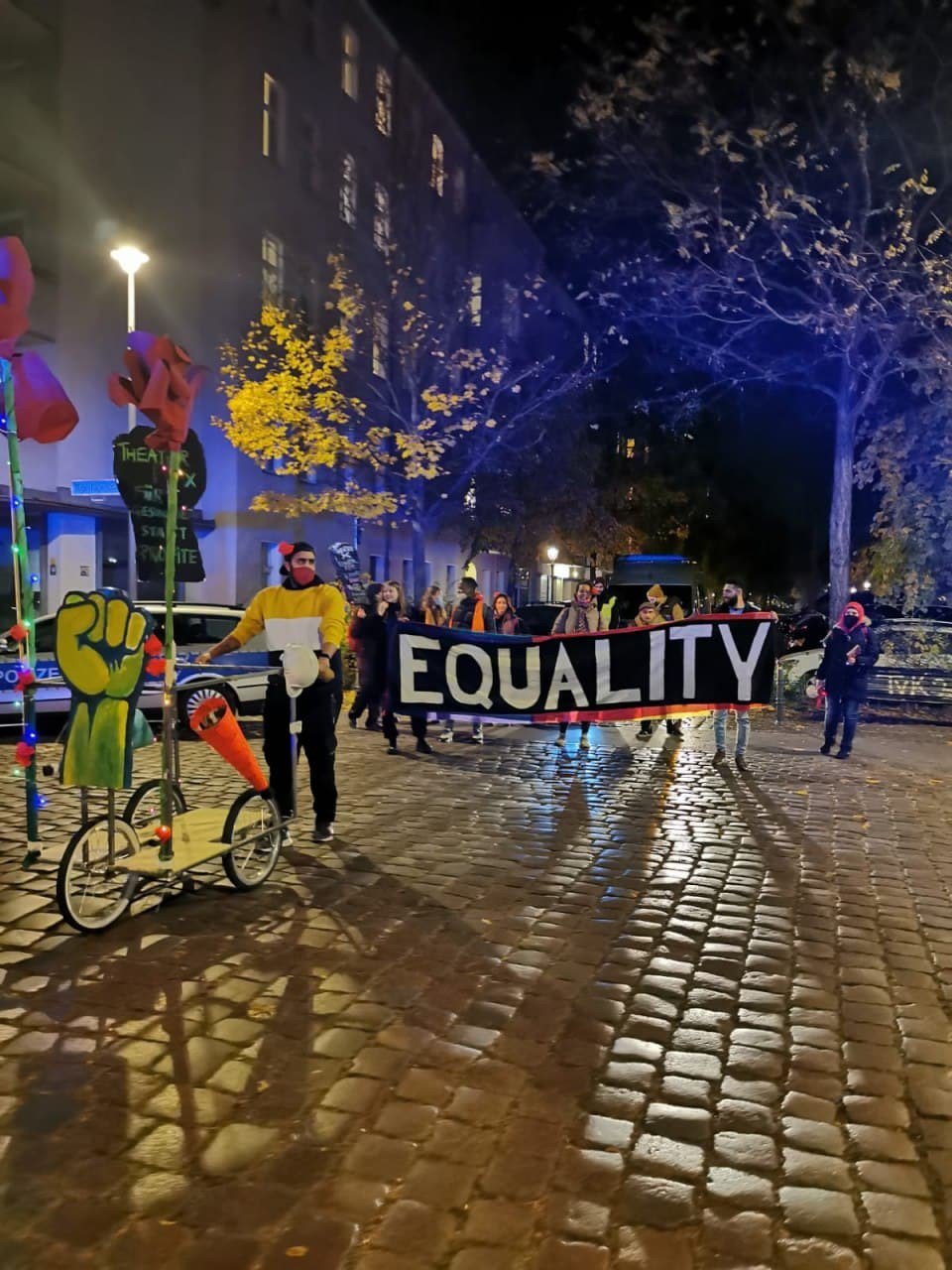 Auf dem Foto von der performativen Kieztour durch Moabit gehen mehrere Personen mit einem Banner und Fahrrädern am Abend. Auf dem Banner steht "Equality". Vor ihnen läuft eine Person mit bunten Papp-Fäusten auf einem Rollwagen.