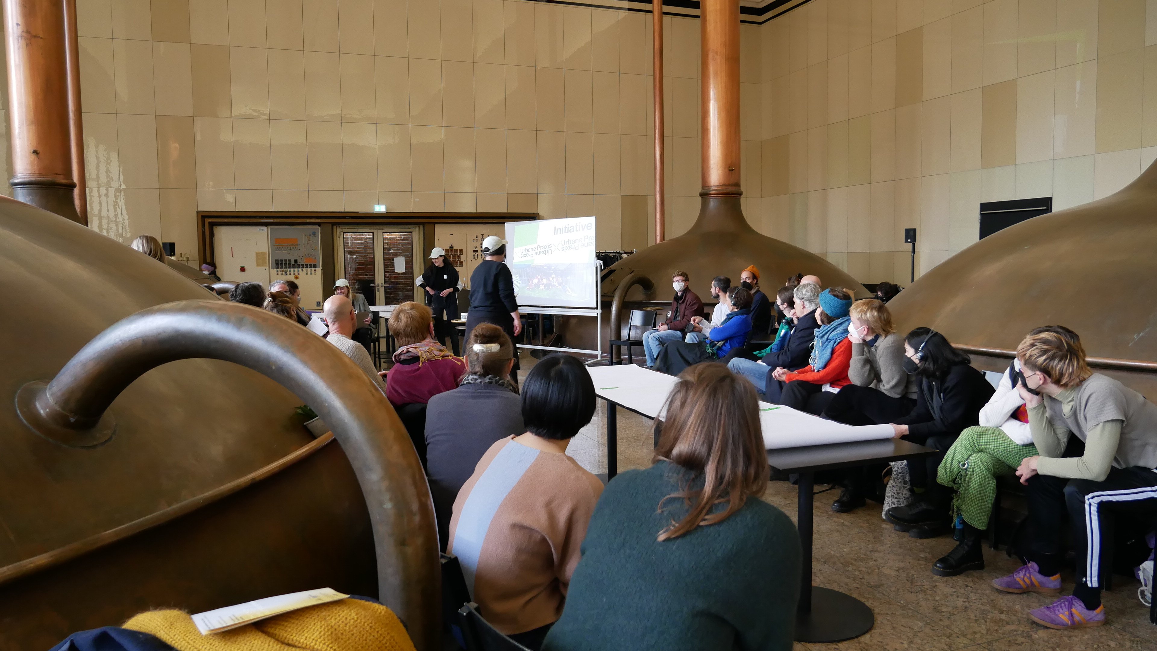 Eine große Gruppe an Menschen sitzt in einem Saal. In der Mitte ist ein Tisch mit drapierten Plakaten, sowie eine Standleinwand. Zwei Personen stehen in der Mitte der Gruppe und geben einen Workshop.