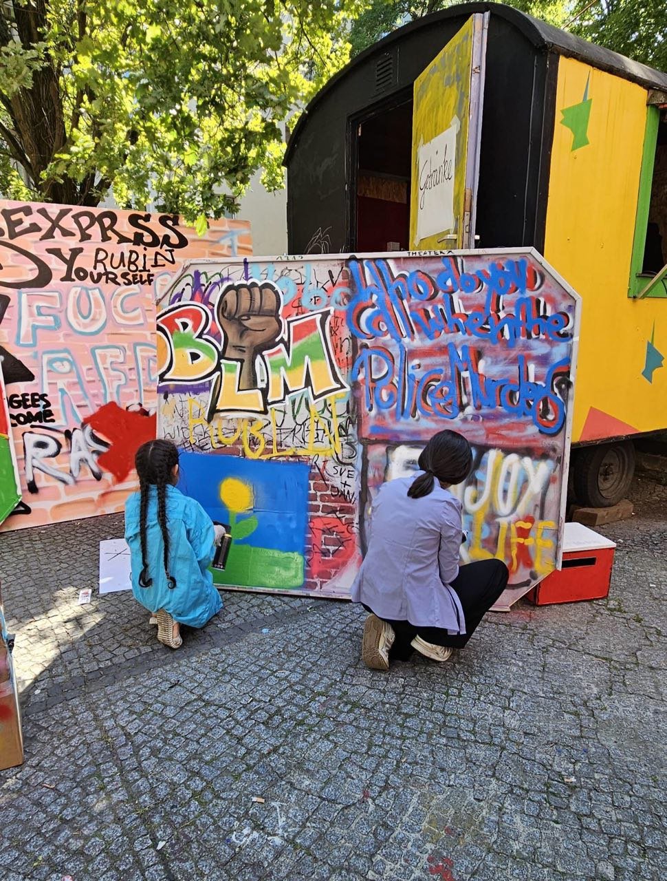 Eine Frau und ein Mädchen sprühen gemeinsam eine Stellwand an. Auf der Stellwand steht: BLM und Who do you call when the police murders.