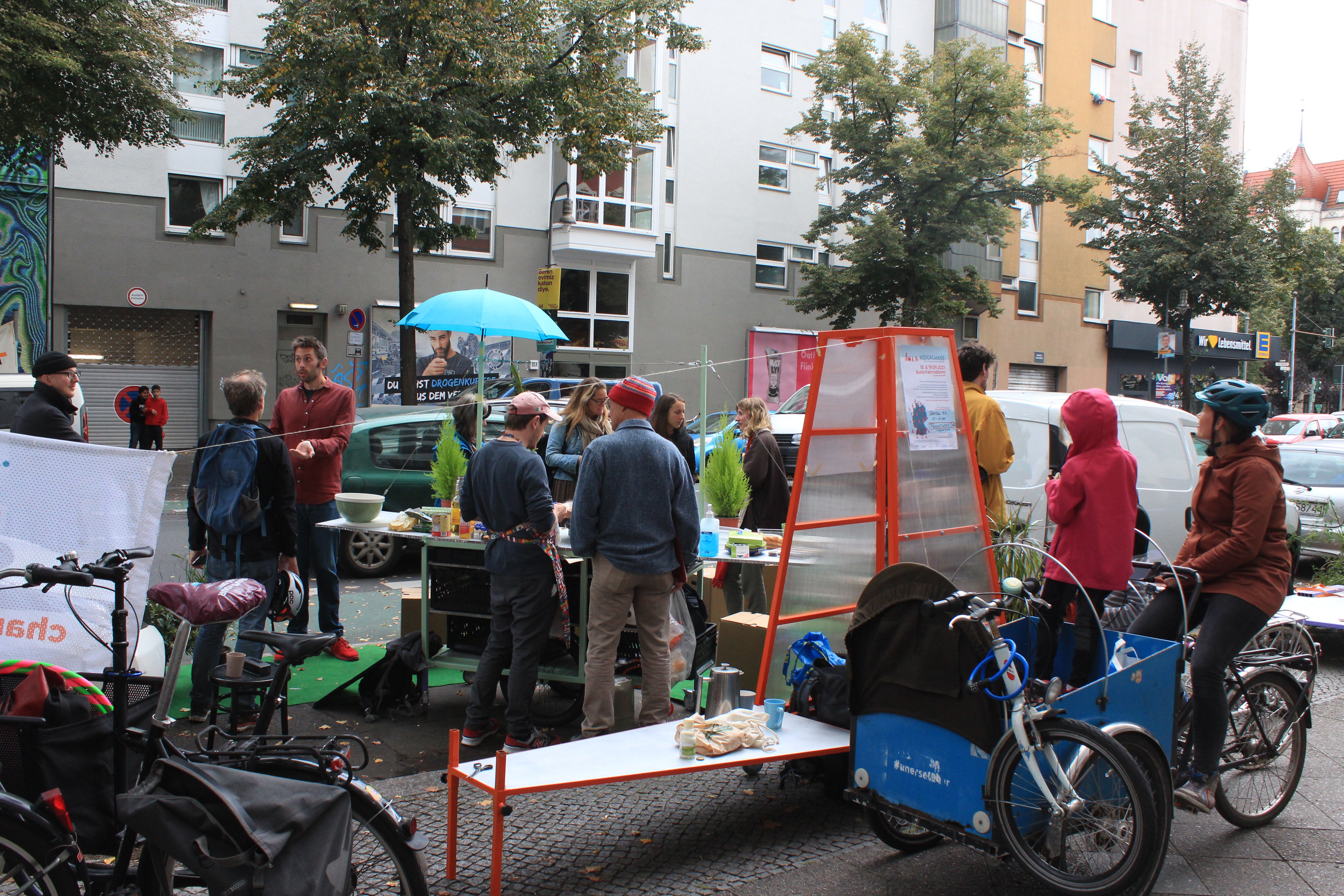 Auf dem Bild sind mehrere Menschen, die auf der Straße stehen zu sehen. Manche von ihnen stehen um einen Tisch, andere unterhalten sich daneben und eine Person sitzt auf dem Fahrrad. Auch andere Fahrräder sind im Vordergrund im Ausschnitt zu sehen.