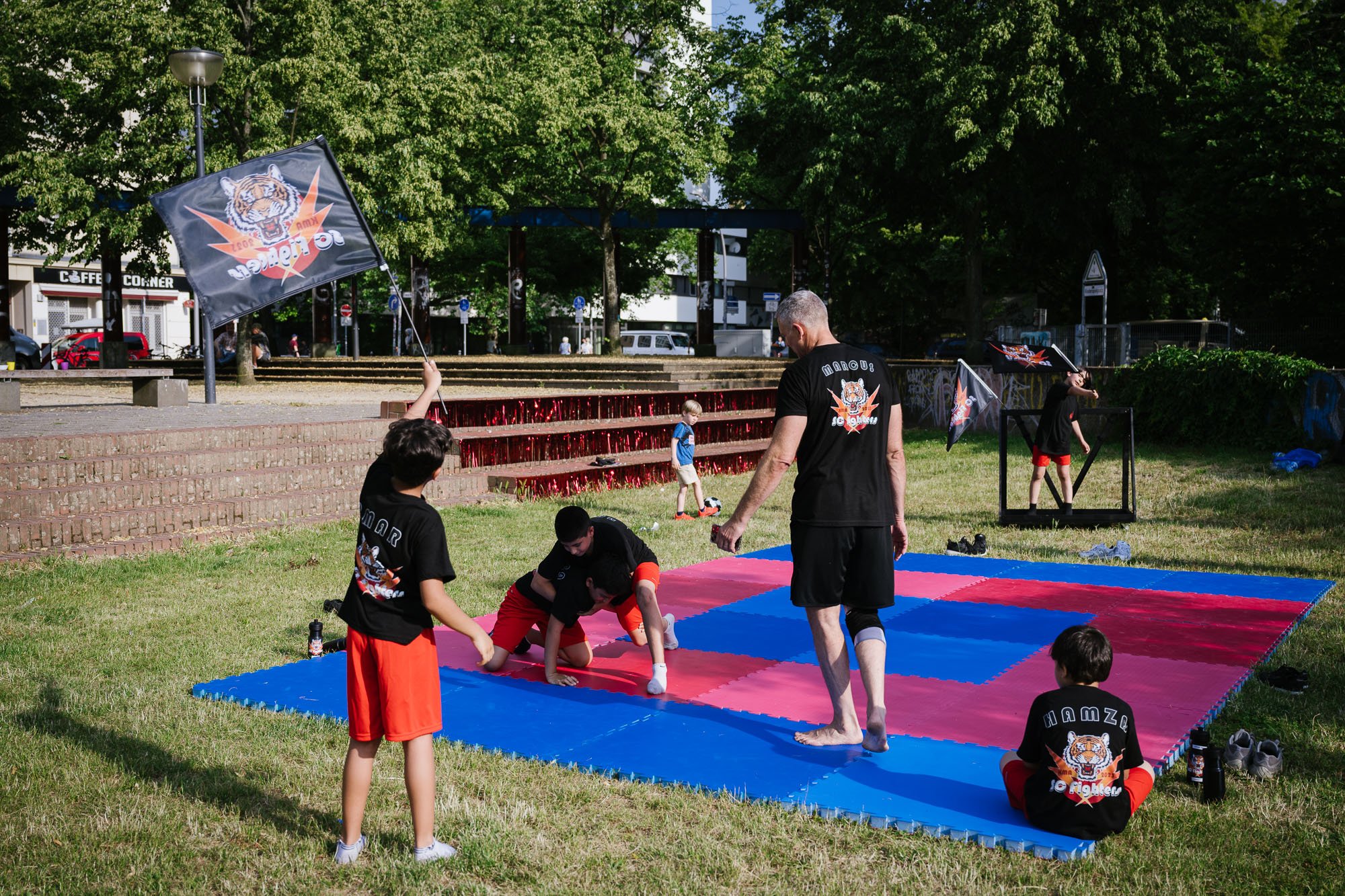 Mehrere Sportmatten sind in einem Park aneinander ausgelegt. Darauf kämpfen zwei Jungen unter Aufsicht eines Trainers. Drumherum sitzen und stehen drei Jungen und schauen zu.