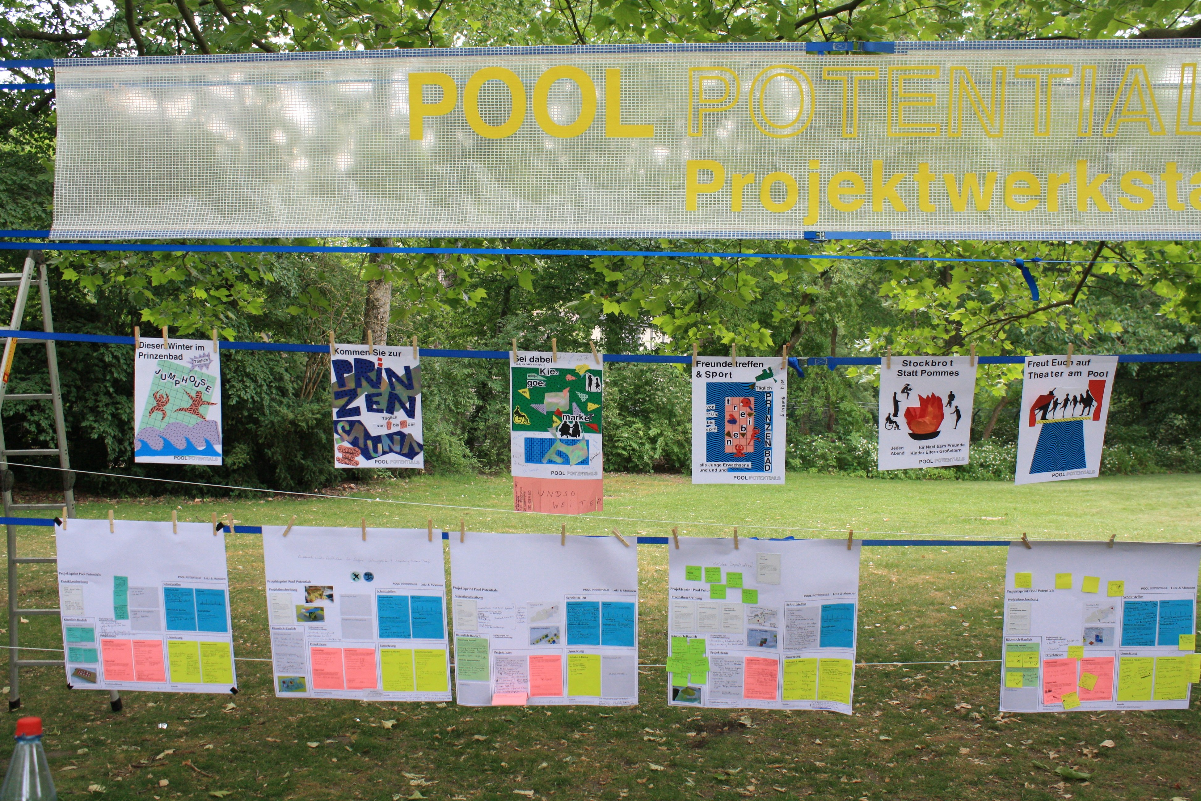 Es sind drei Leinen in einem Park gespannt worden. Auf den Leinen hängt ganz oben eine Plane mit der Überschrift "Pool Potentials Projekt". Darunter sind verschiedene Flyer angeheftet. Und darunter sind Blätter mit kleinen Zetteln angeheftet.
